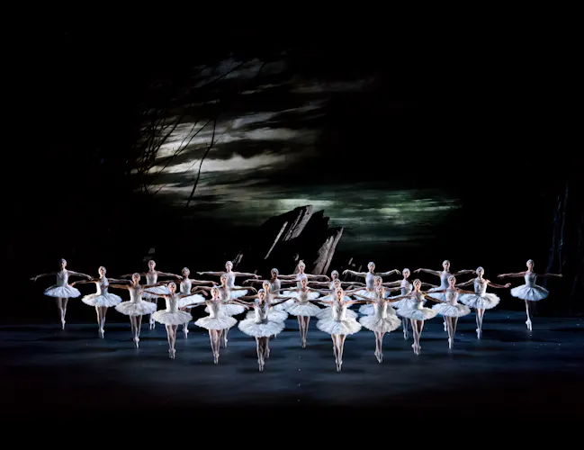 A view from above onto a dark set stage with several ballet dancers wearing white tutus and white feathered headdresses standing en pointe in a triangle shaped formation. They perform in front of a dark rock front and the background is dark, moody skies. They are performing in The Royal Ballet's production of Swan Lake. (1 of 5)