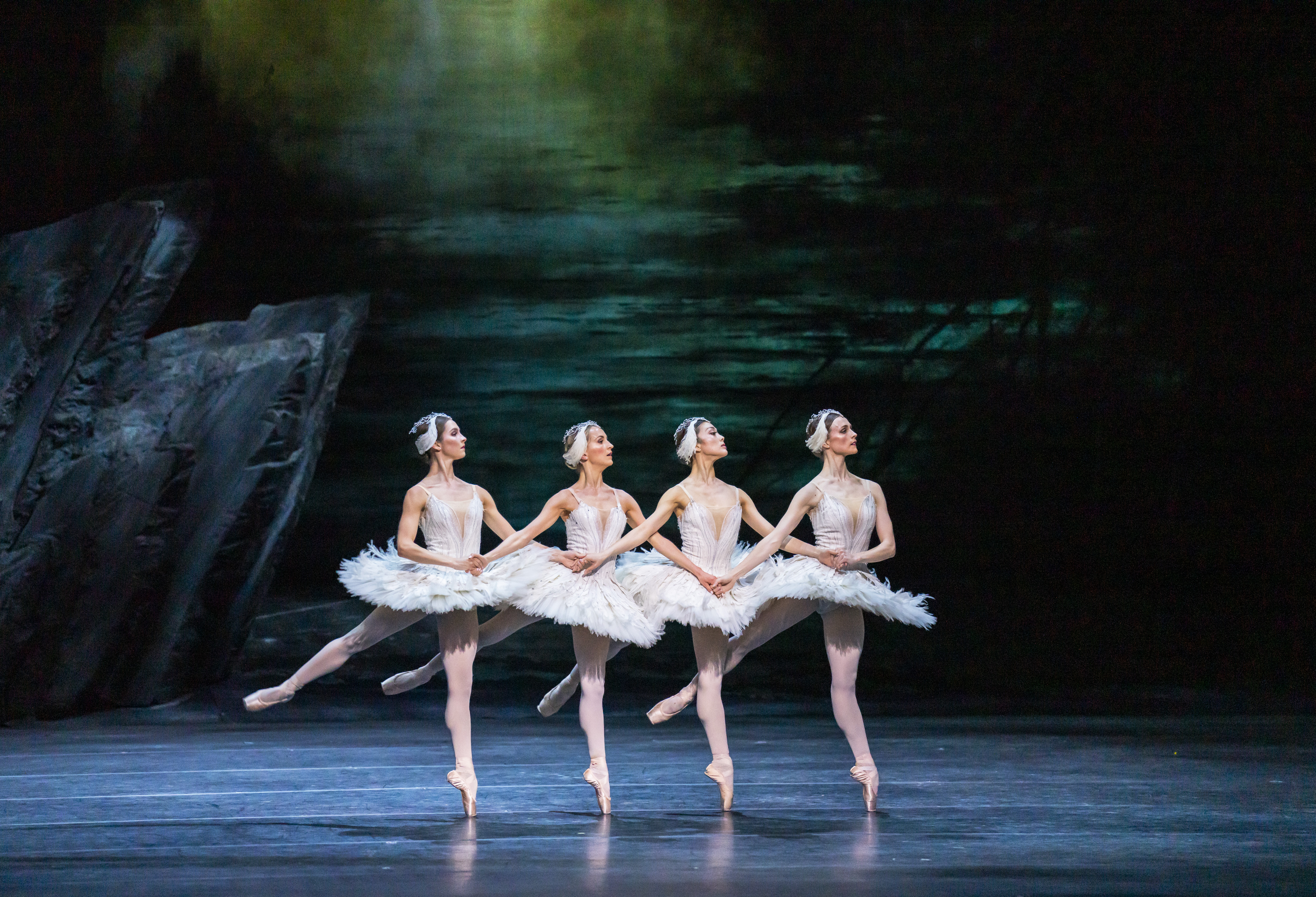Four ballet dancers wearing white tutus and white feather headdresses perform together on stage in front of a moody sky backdrop and a sharp rock face. They are performing as swans in The Royal Ballet's production of Swan Lake. (3 of 5)