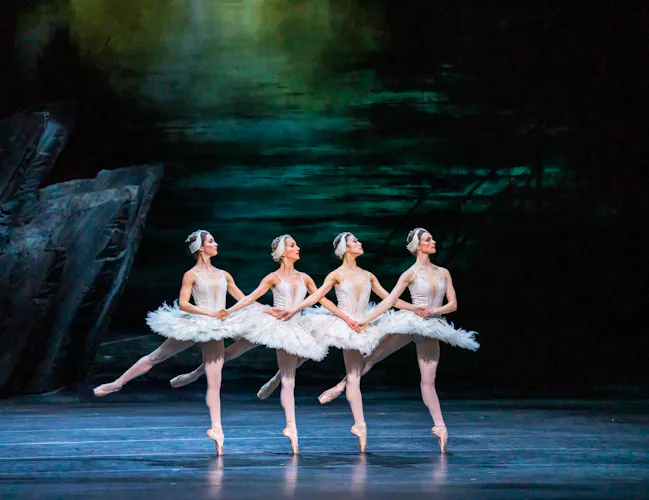 Four ballet dancers wearing white tutus and white feather headdresses perform together on stage in front of a moody sky backdrop and a sharp rock face. They are performing as swans in The Royal Ballet's production of Swan Lake. (3 of 5)