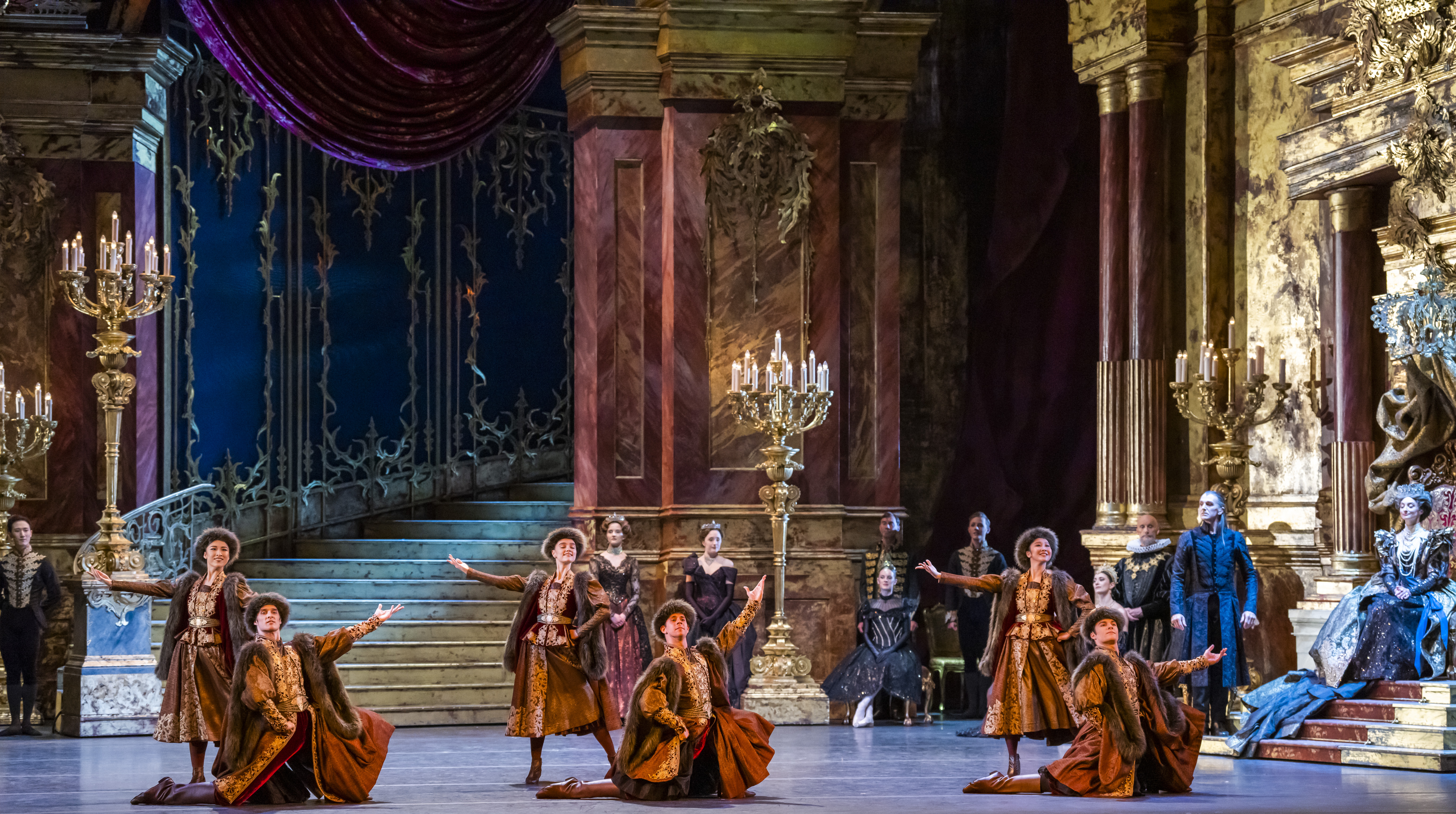 Several ballet dancers in traditional Russian costumes perform on stage that is set like an ornate ballroom with a curved staircase. (4 of 5)