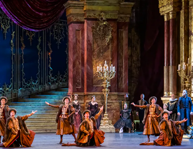 Several ballet dancers in traditional Russian costumes perform on stage that is set like an ornate ballroom with a curved staircase. (4 of 5)