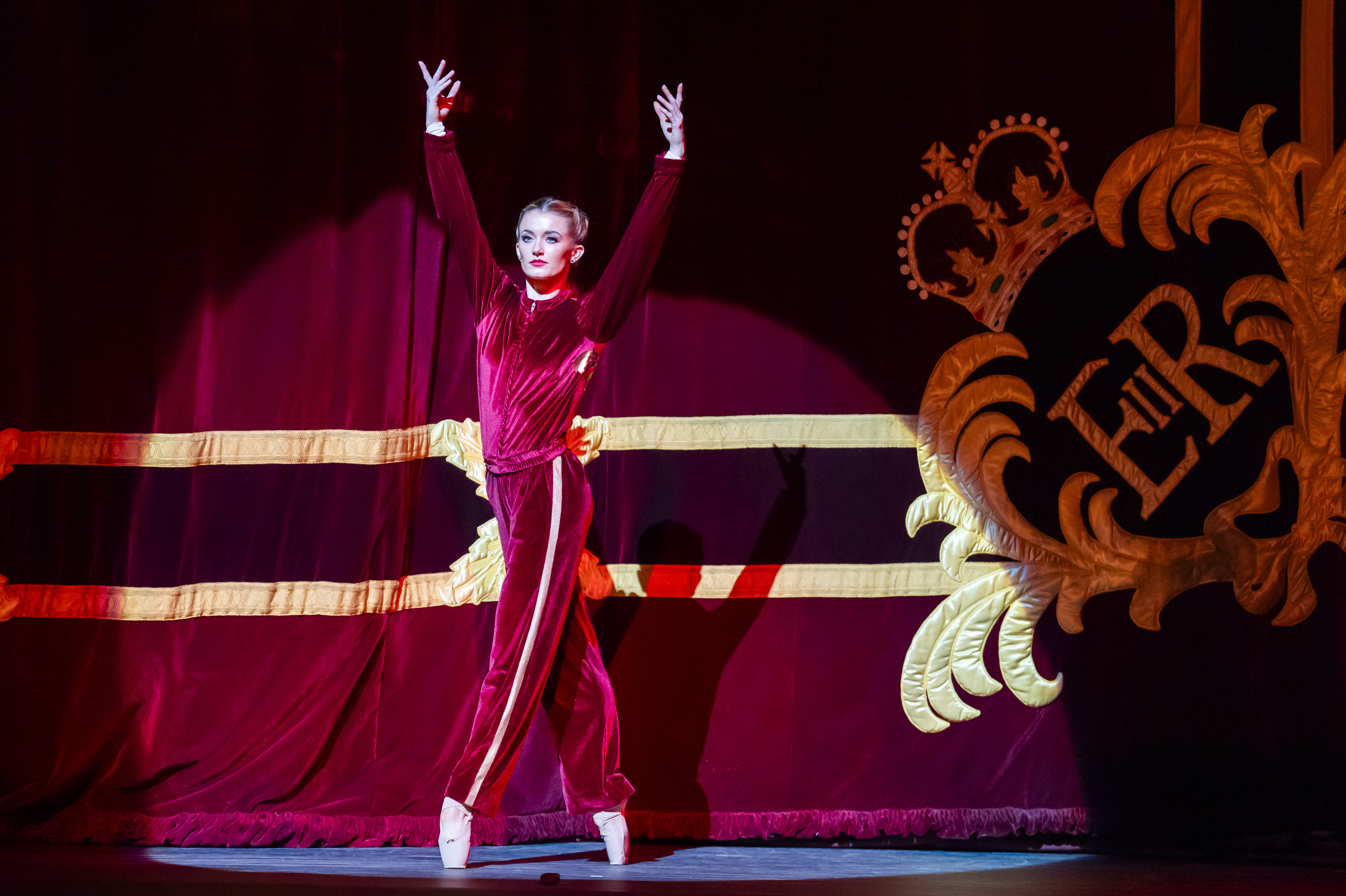 The Royal Ballet dancer Anna Rose O'Sullivan stands en pointe wearing a red velvet tracksuit jacket and trousers in front of the red and gold embroidered curtain of the Royal Opera House. (1 of 8)