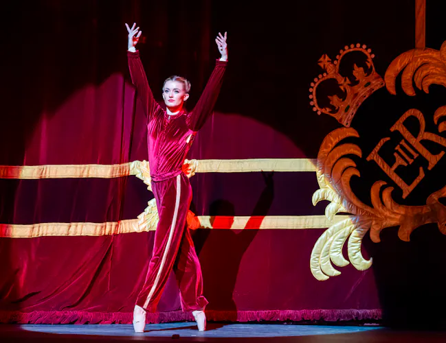 The Royal Ballet dancer Anna Rose O'Sullivan stands en pointe wearing a red velvet tracksuit jacket and trousers in front of the red and gold embroidered curtain of the Royal Opera House. (1 of 8)