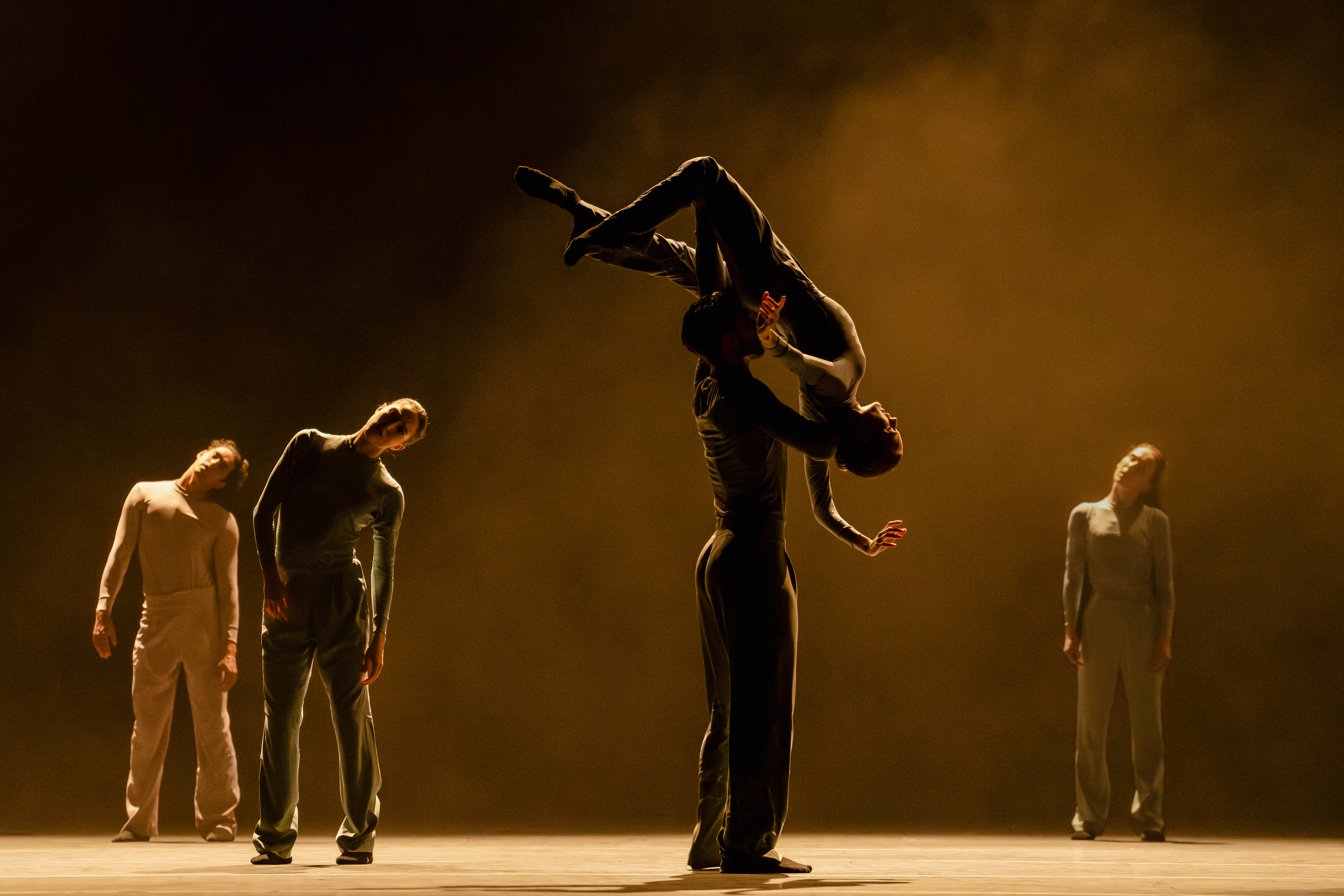 A ballet dancer holds another dancer high above their shoulders with their head and arms draped downwards. They are flanked by three other dancers behind them. The stage is stark with a haze. 