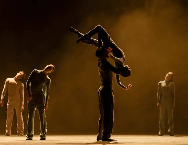 A ballet dancer holds another dancer high above their shoulders with their head and arms draped downwards. They are flanked by three other dancers behind them. The stage is stark with a haze. (7 of 8)