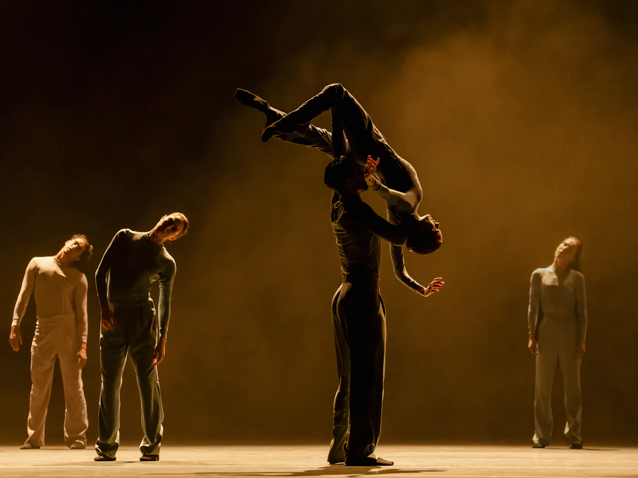 A ballet dancer holds another dancer high above their shoulders with their head and arms draped downwards. They are flanked by three other dancers behind them. The stage is stark with a haze.