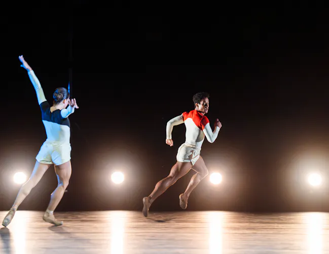 Two ballet dancers wearing monochrome costumes chase one another on a stark stage with bright lights facing the audience. (4 of 8)