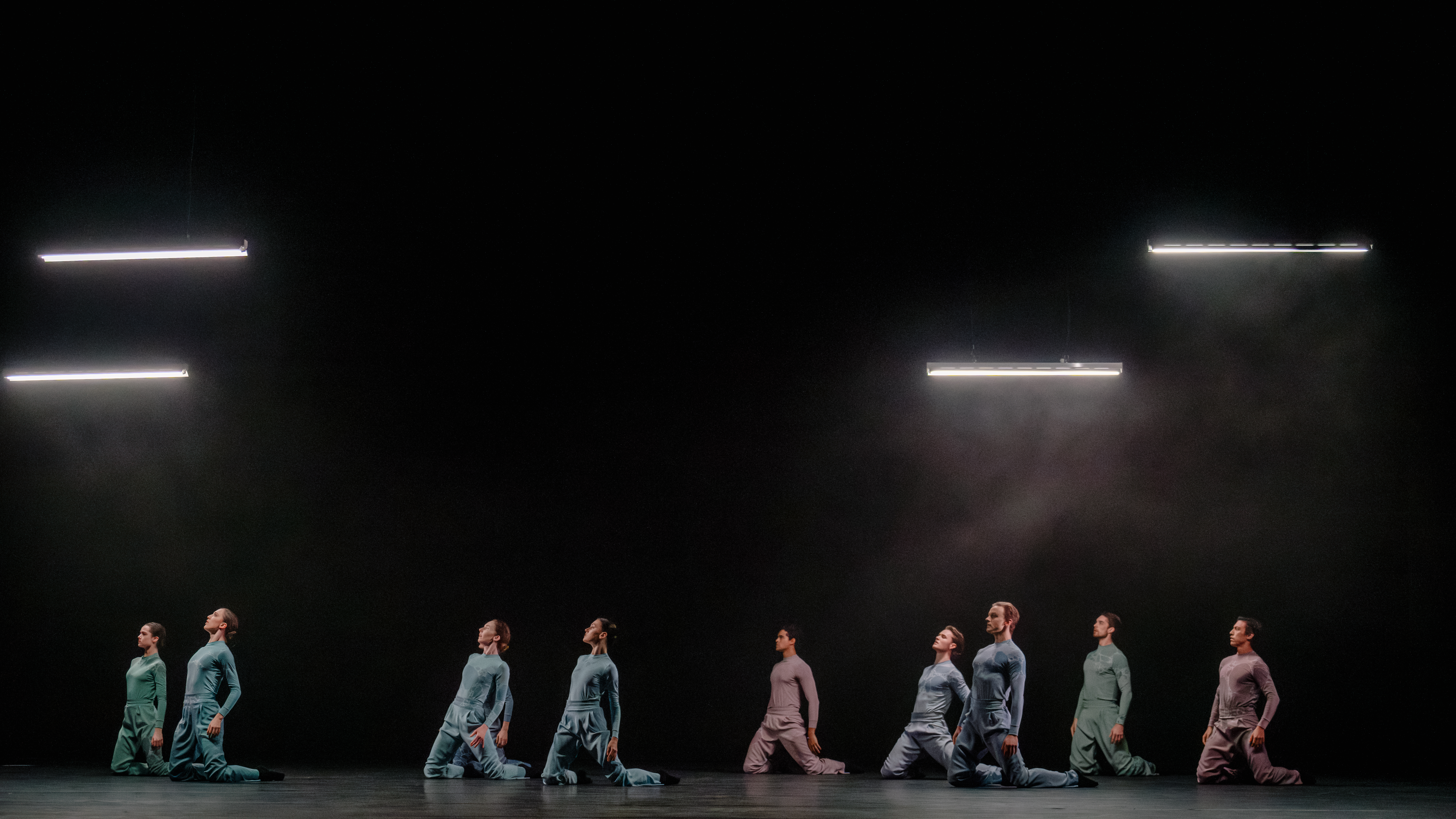 Several ballet dancers sit on their knees wearing jumpsuits. They are against a black backdrop with four neon striplights above them. (8 of 8)