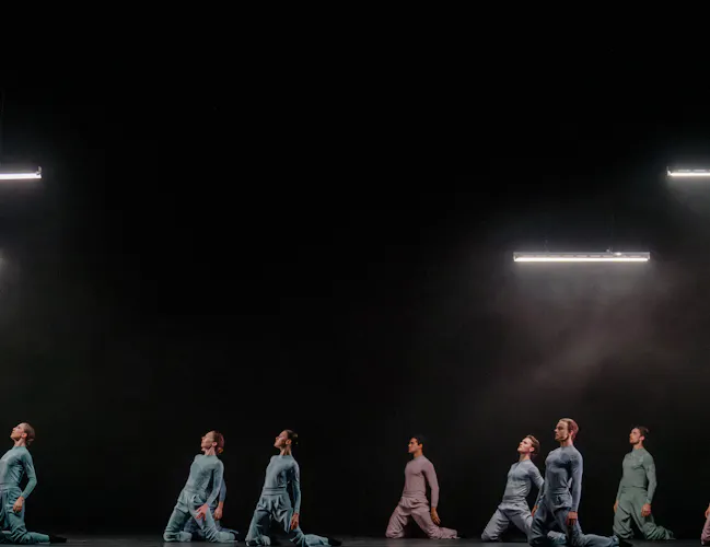 Several ballet dancers sit on their knees wearing jumpsuits. They are against a black backdrop with four neon striplights above them. (8 of 8)