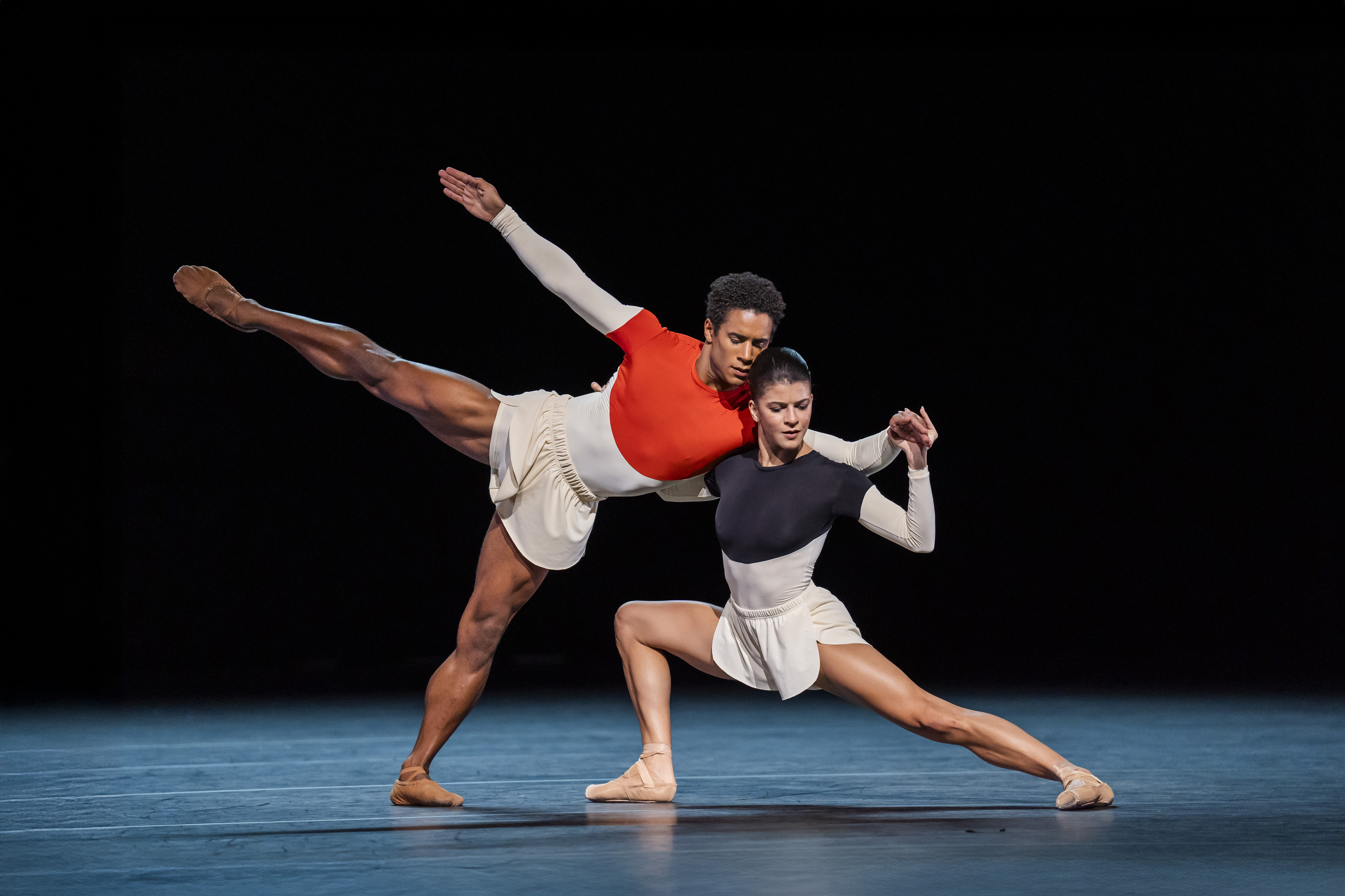 Two Royal Ballet dancers perform on stage wearing monochrome leotards and white shorts. One dancer is in a lunge position supporting the other dancer who leans against them. The stage is stark with a dark backdrop. 