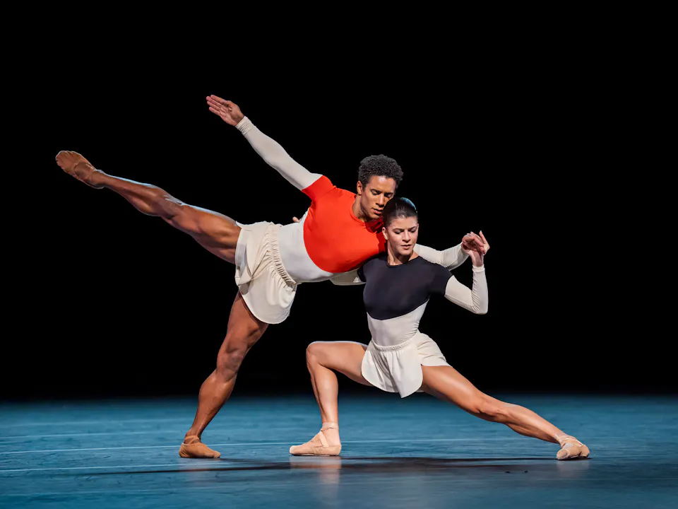 Two Royal Ballet dancers perform on stage wearing monochrome leotards and white shorts. One dancer is in a lunge position supporting the other dancer who leans against them. The stage is stark with a dark backdrop.