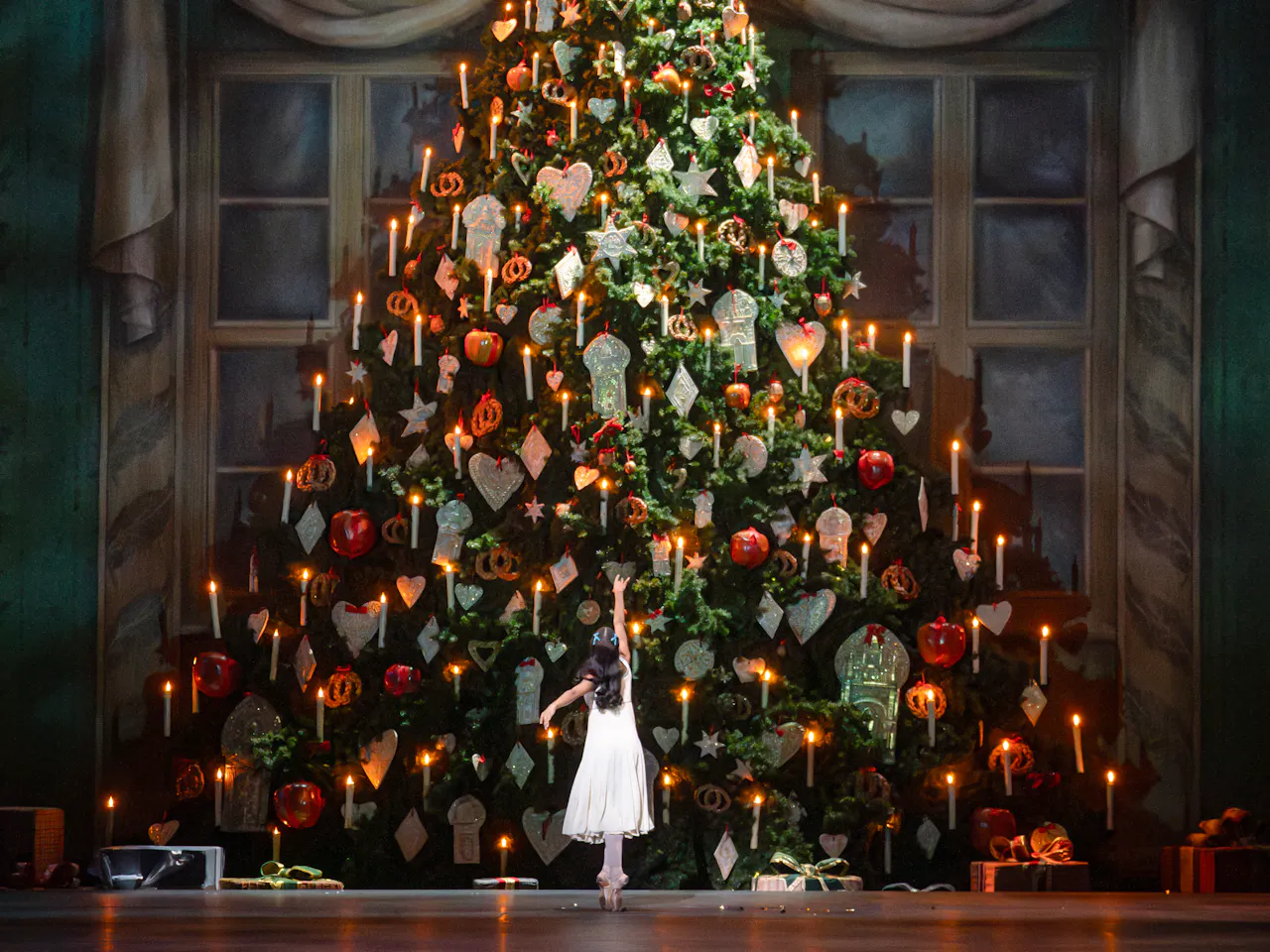 A ballet dancer wearing a white dress stands with their back to the audience looking up at an enormously, luxuriously dressed Christmas tree with presents below that sits in front of a backdrop painted as large dressed windows. It is a scene from The Royal Ballet's production of The Nutcracker.