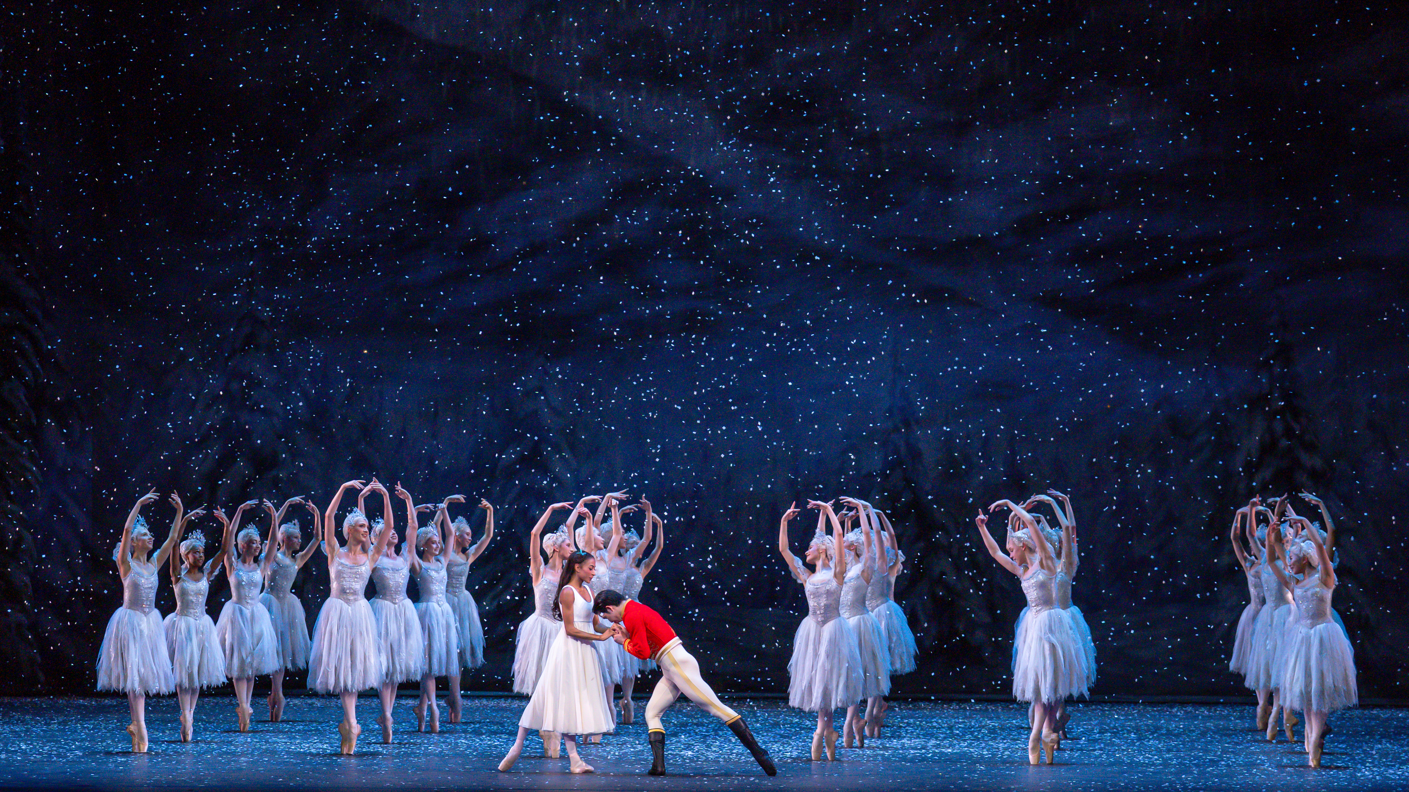 A ballet dancer wearing a red jacket holds the hands of another ballet dancer wearing a white dress on a stage set like a snowy night. Behind them are a group of ballet dancers wearing white tutus, en pointe in a formation with their arms raised above their heads. They are performing in The Royal Ballet's production of The Nutcracker. (5 of 5)