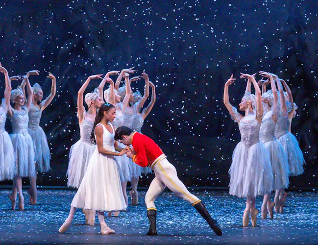 A ballet dancer wearing a red jacket holds the hands of another ballet dancer wearing a white dress on a stage set like a snowy night. Behind them are a group of ballet dancers wearing white tutus, en pointe in a formation with their arms raised above their heads. They are performing in The Royal Ballet's production of The Nutcracker. (5 of 5)