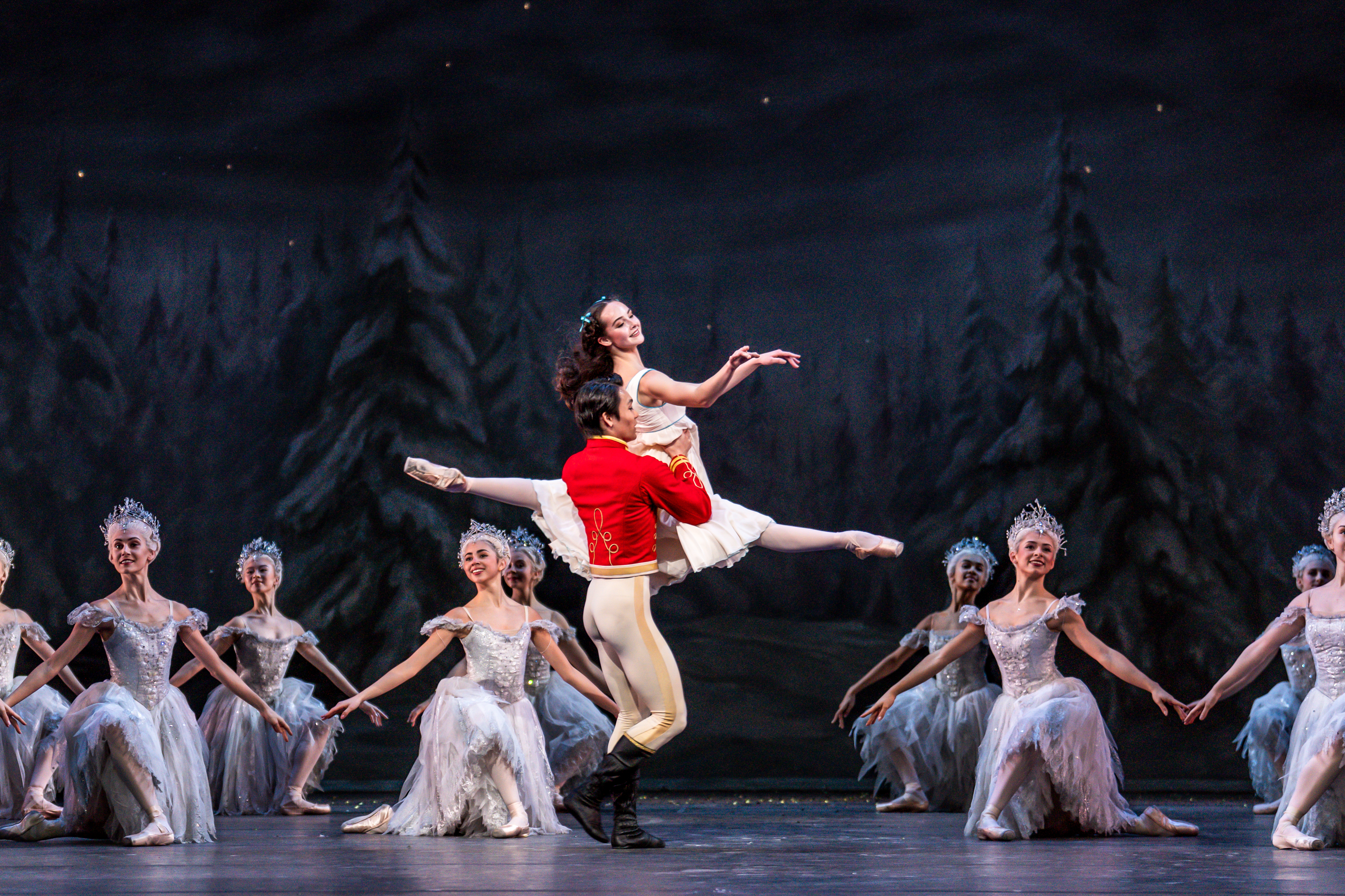 A ballet dancer wearing a red military jacket holds the waist of a ballet dancer wearing a white dress as she launches herself into the air with her legs stretched vertically. Behind them ballet dancers wearing white tutus and headdresses are in formation on one knee. They are all performing on stage with a backdrop of tall fir snow-flecked fir trees. They are performing in The Royal Ballet's production of The Nutcracker. (4 of 5)
