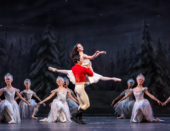 A ballet dancer wearing a red military jacket holds the waist of a ballet dancer wearing a white dress as she launches herself into the air with her legs stretched vertically. Behind them ballet dancers wearing white tutus and headdresses are in formation on one knee. They are all performing on stage with a backdrop of tall fir snow-flecked fir trees. They are performing in The Royal Ballet's production of The Nutcracker. (4 of 5)