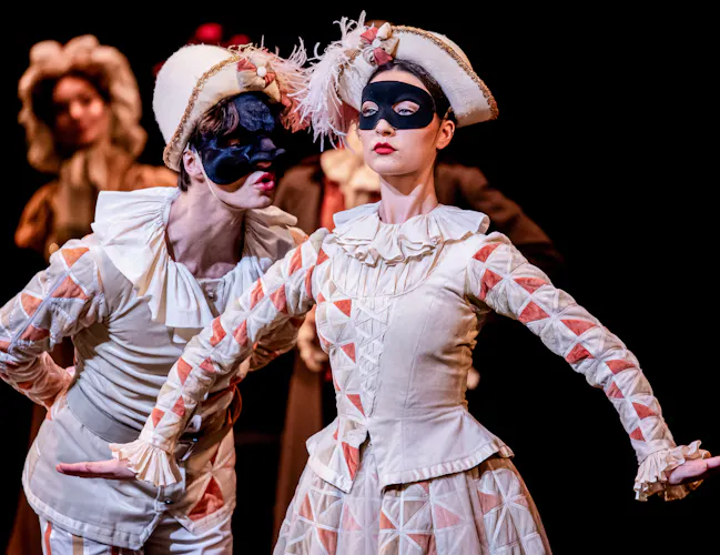 Two ballet dancers wearing a harlequin costumes, feather capped hats and black eye masks perform on stage. It is a scene from The Royal Opera House in The Royal Ballet's production of The Nutcracker. (2 of 5)