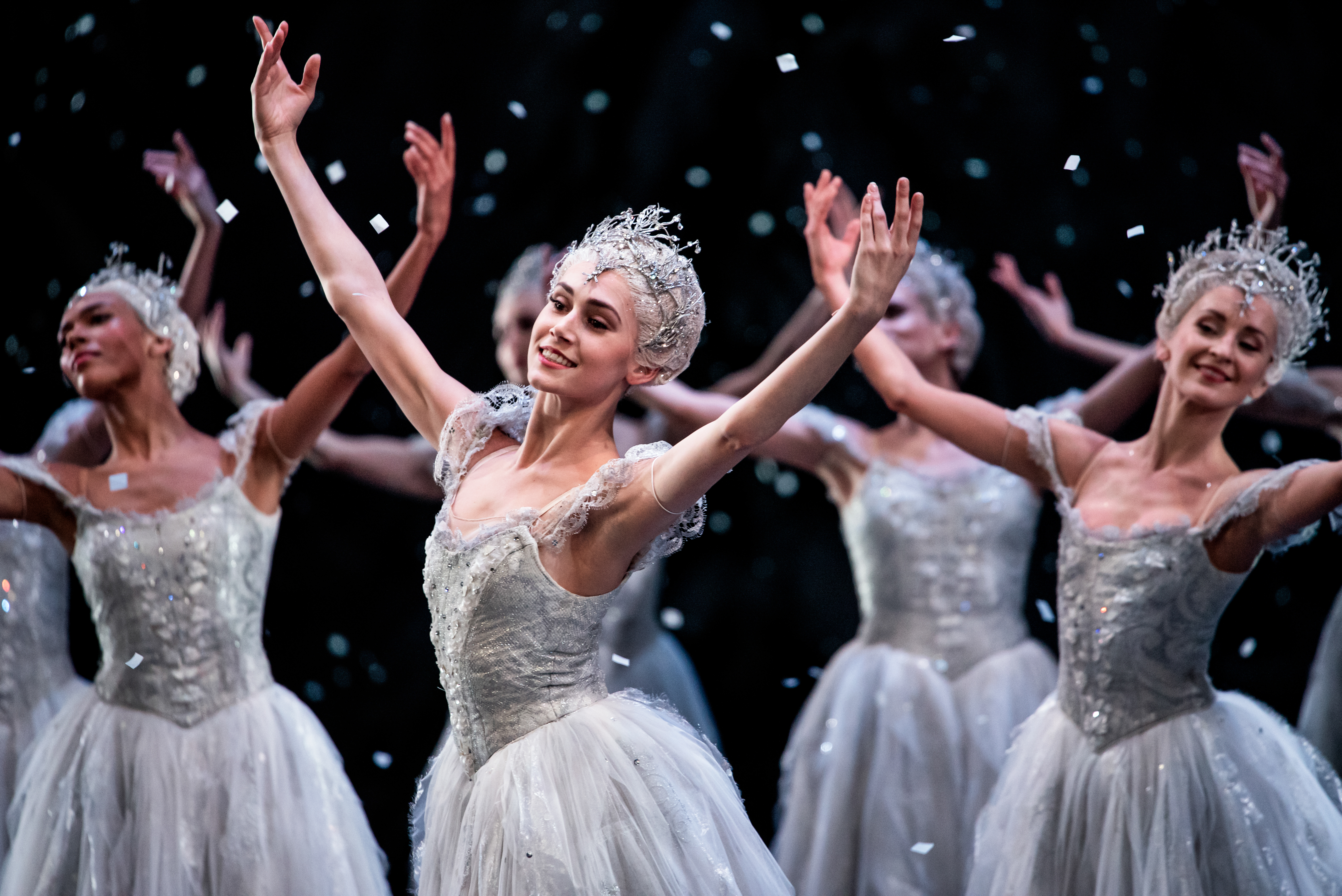 Ballet dancers wearing white tutus and elaborate tiara headdresses stand on stage with their arms raised above. They are performing as snowflakes in The Royal Ballet's production of The Nutcracker. (3 of 5)