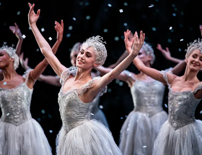 Ballet dancers wearing white tutus and elaborate tiara headdresses stand on stage with their arms raised above. They are performing as snowflakes in The Royal Ballet's production of The Nutcracker. (3 of 5)