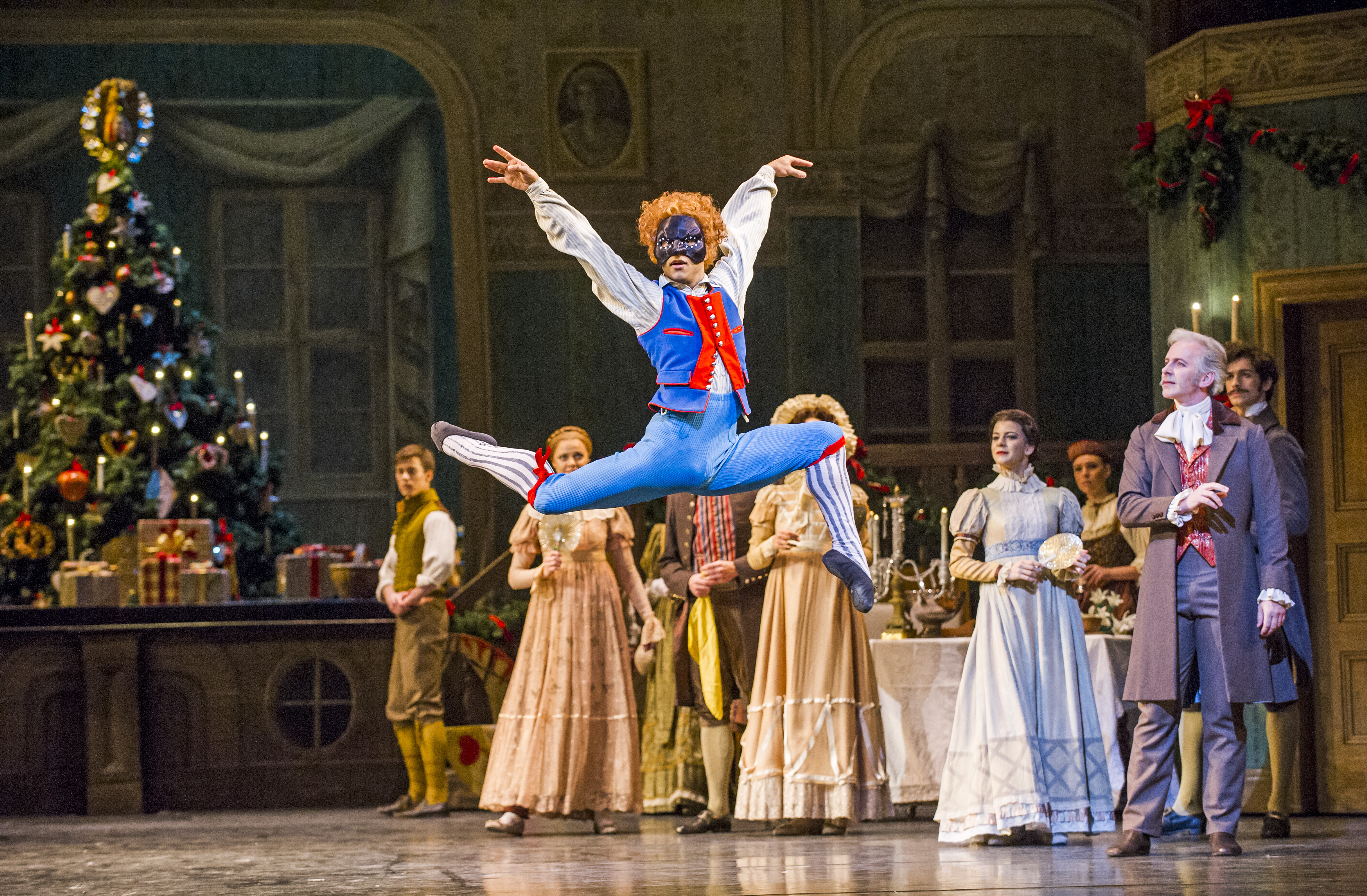 A ballet dancer wearing blue breeches, patterned long socks, a blue and red waistcoat and black harlequin face mask launches themselves into the air on stage. Behind them the scene is set like an extravagant Christmas party. They are performing in The Royal Ballet's production of The Nutcracker. (1 of 5)