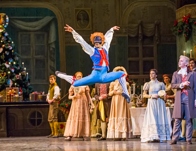 A ballet dancer wearing blue breeches, patterned long socks, a blue and red waistcoat and black harlequin face mask launches themselves into the air on stage. Behind them the scene is set like an extravagant Christmas party. They are performing in The Royal Ballet's production of The Nutcracker. (1 of 5)