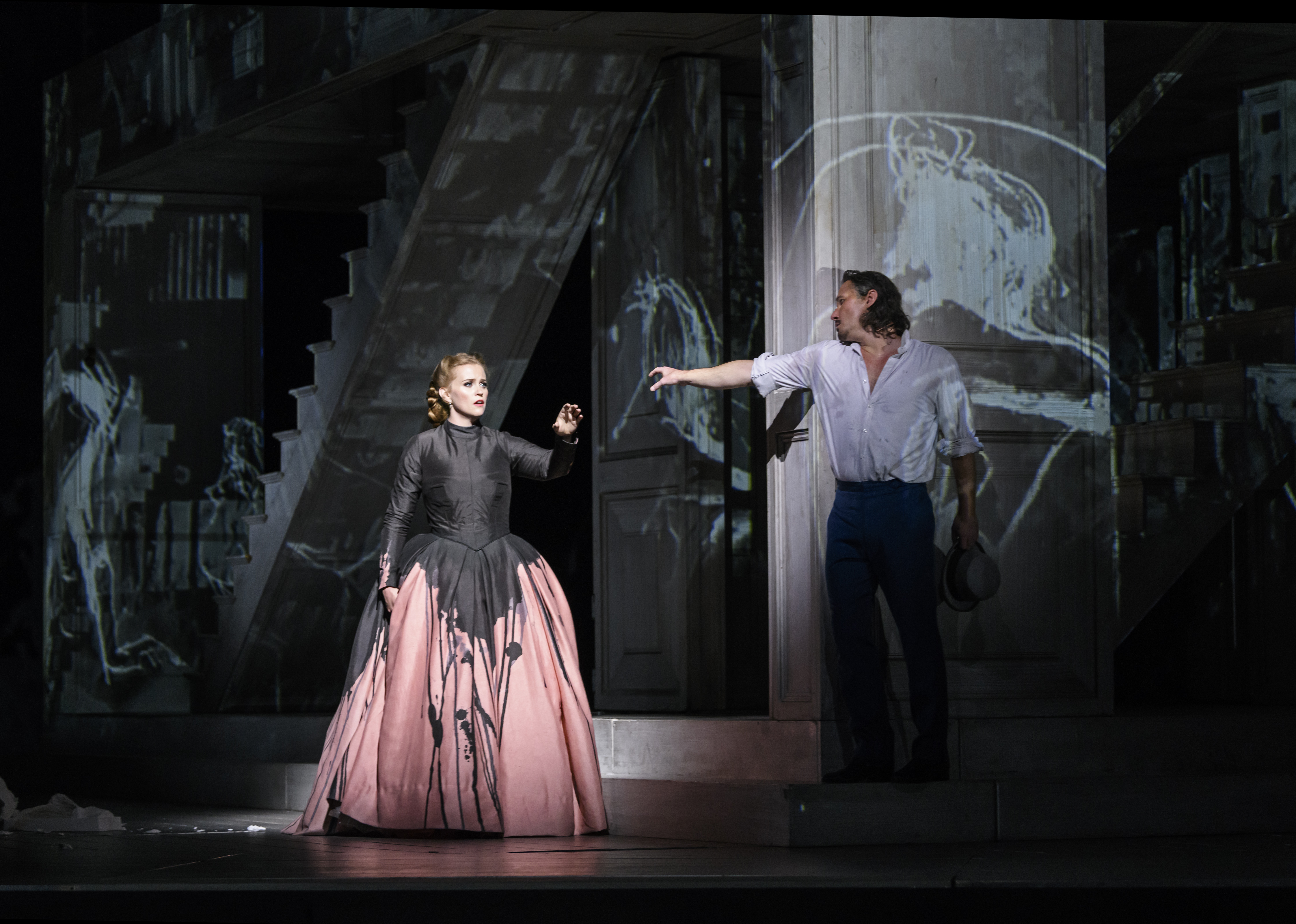 Two opera singers stand on stage. One wears a pink and black ballgown and holds her arm out towards the other singer who wears a white shirt. They are performing in The Royal Opera's production of Don Giovanni. (4 of 4)