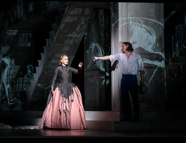 Two opera singers stand on stage. One wears a pink and black ballgown and holds her arm out towards the other singer who wears a white shirt. They are performing in The Royal Opera's production of Don Giovanni. (4 of 4)