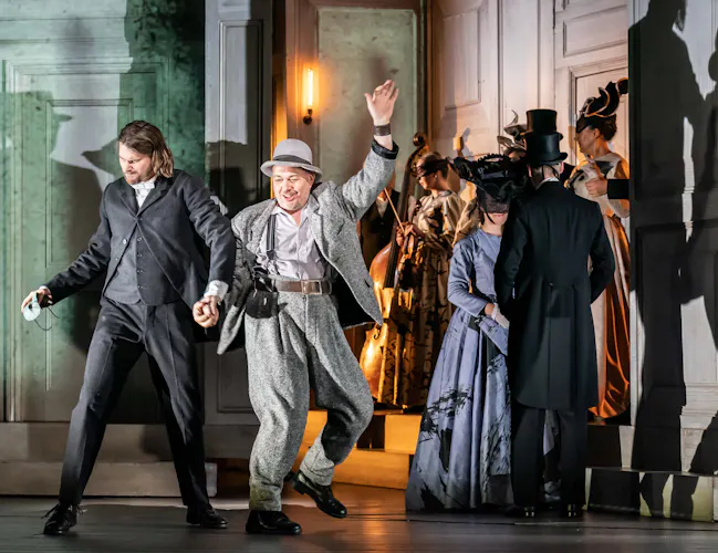 Several performers stand around a party scene in a traditionally decorated room on stage of The Royal Opera House in The Royal Opera's production of Don Giovanni. (3 of 4)