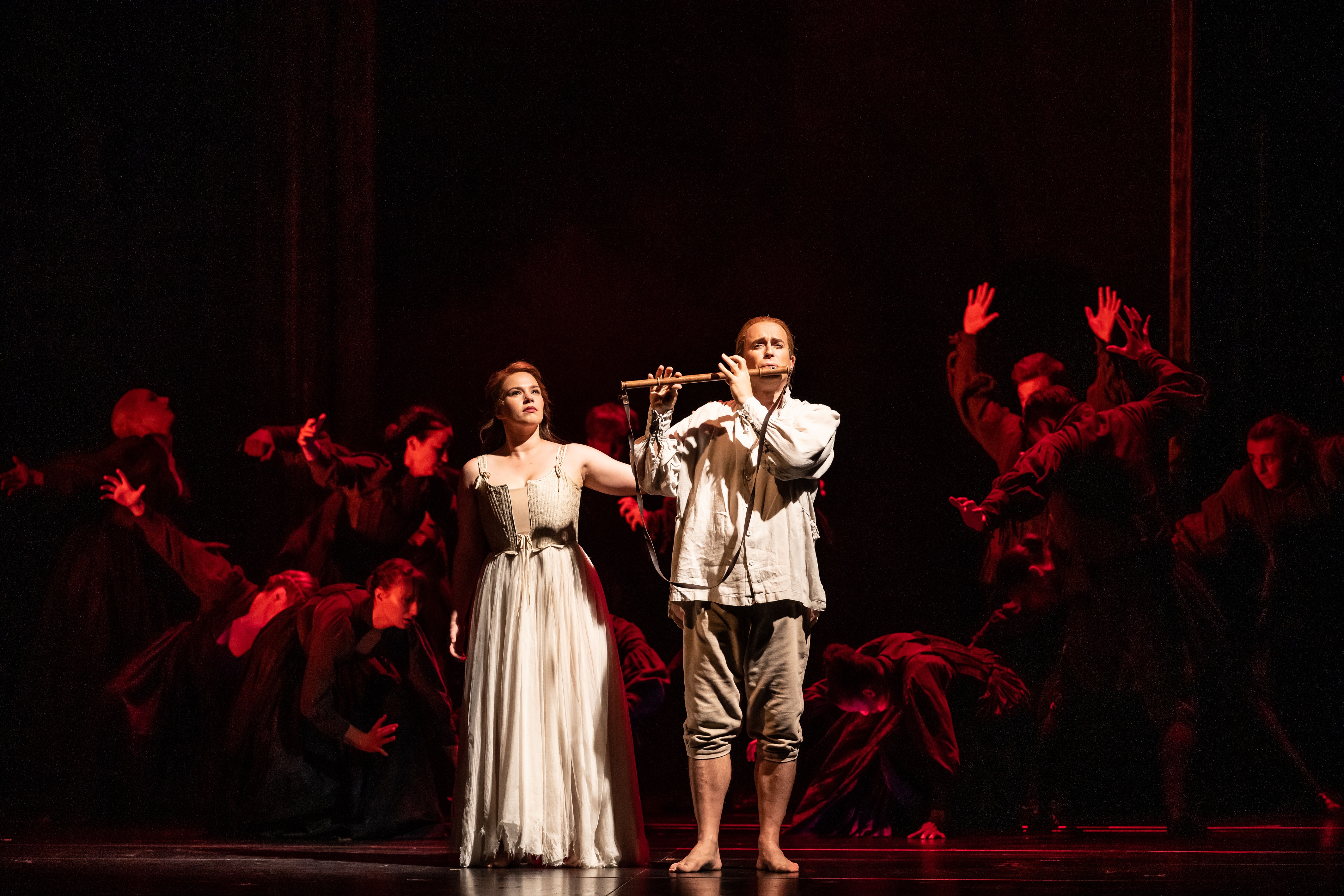 Two performers stand on stage of The Royal Opera House. One holds a flute up and plays. They are lit brightly but behind them other performers are made visible in the darkness with red lighting. They are performing in The Royal Opera's production of The Magic Flute. (1 of 7)