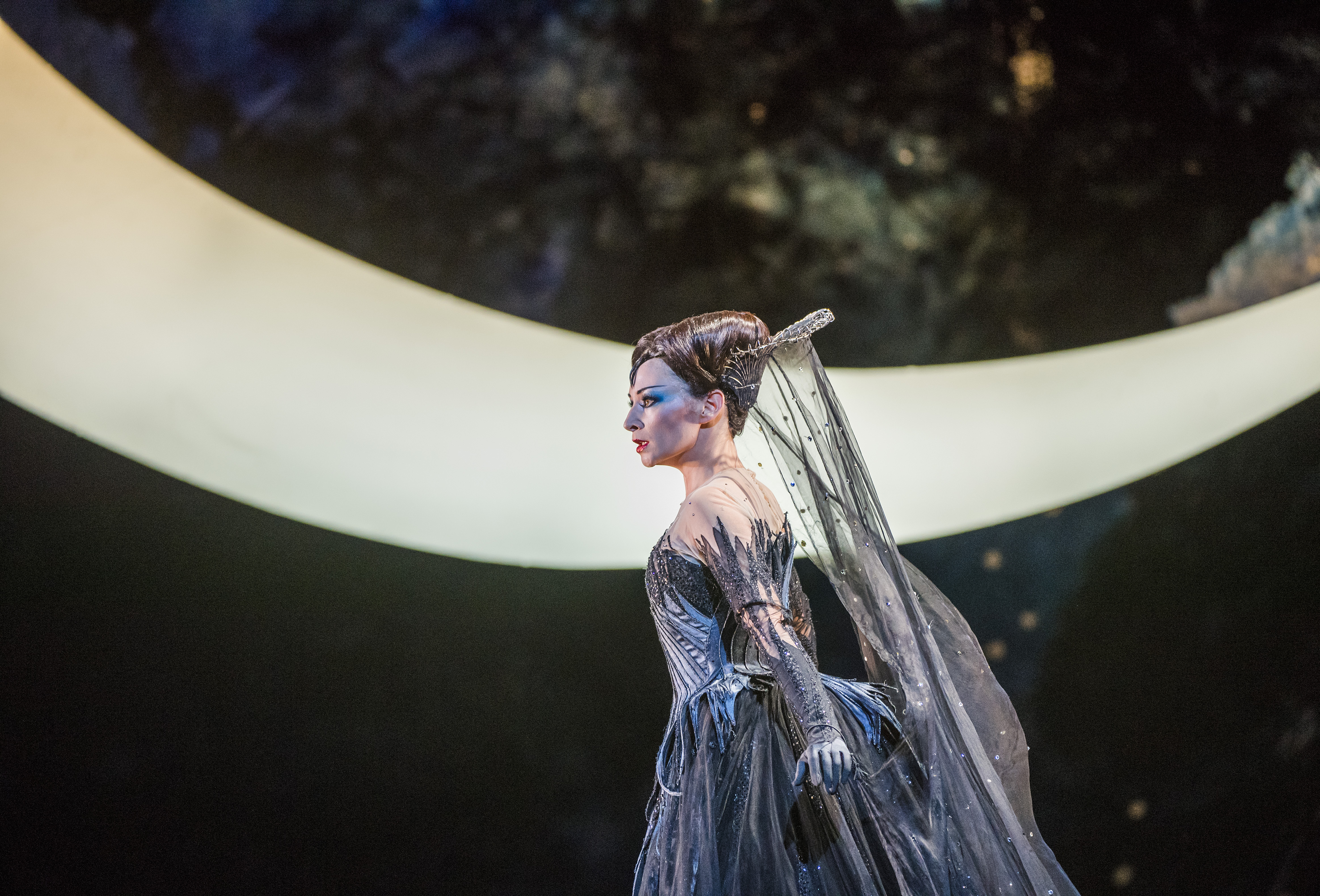 An opera singer wearing a grey dress and medieval-style headdress stands on stage in front of a large lit moon backdrop. They are performing in The Royal Opera's production of The Magic Flute. (2 of 7)