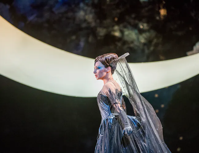 An opera singer wearing a grey dress and medieval-style headdress stands on stage in front of a large lit moon backdrop. They are performing in The Royal Opera's production of The Magic Flute. (2 of 7)