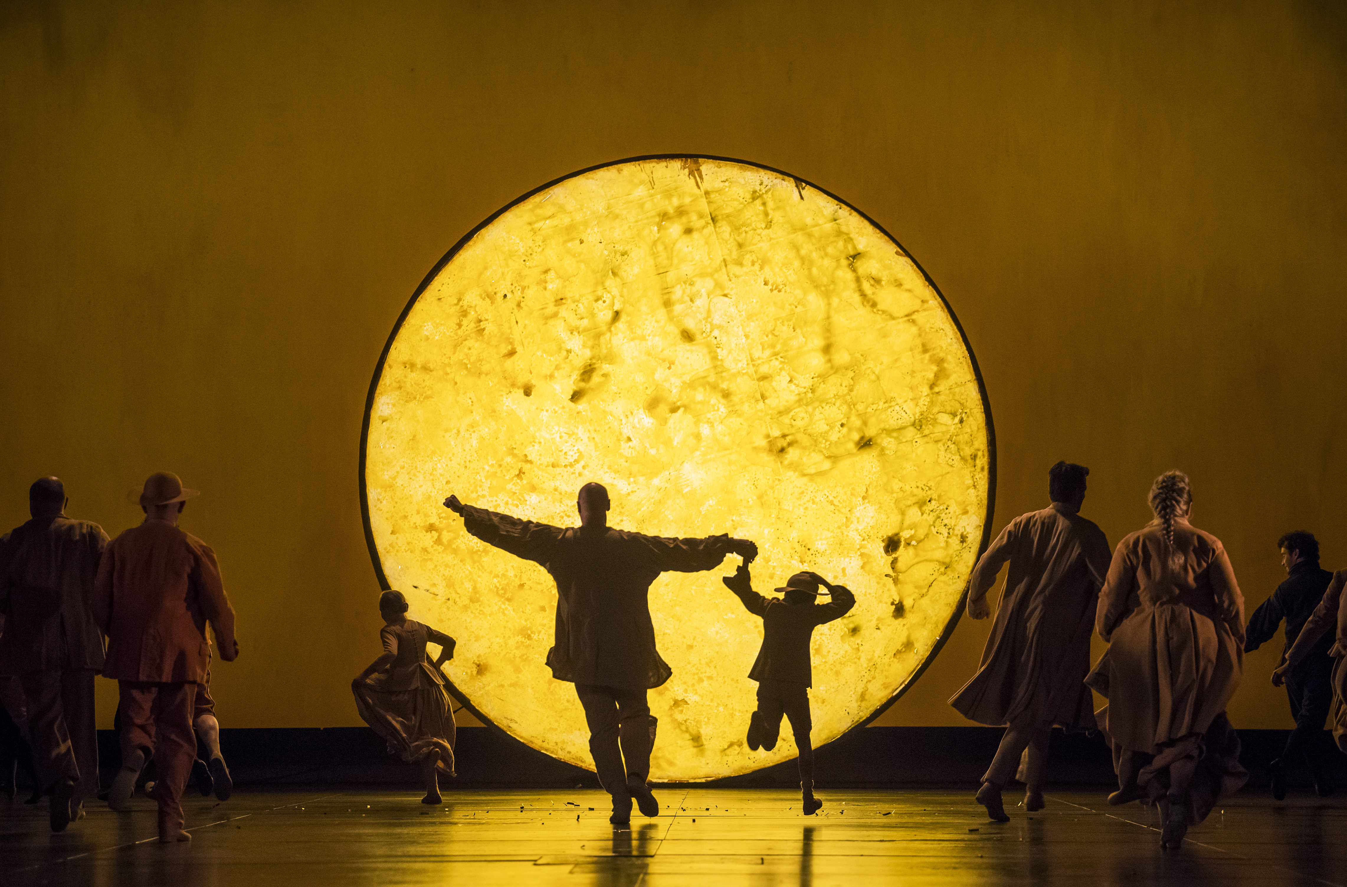 A brightly lit yellow light painted to look like the surface of a star or planet is in the centre of the stage. An adult and child run towards the light with their back to the audience while other members of the chorus run alongside. They are performing in The Royal Opera's production of The Magic Flute. (3 of 7)