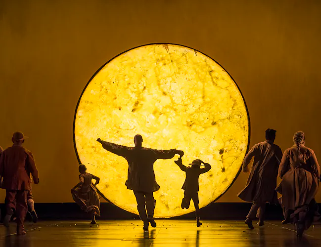 A brightly lit yellow light painted to look like the surface of a star or planet is in the centre of the stage. An adult and child run towards the light with their back to the audience while other members of the chorus run alongside. They are performing in The Royal Opera's production of The Magic Flute. (3 of 7)
