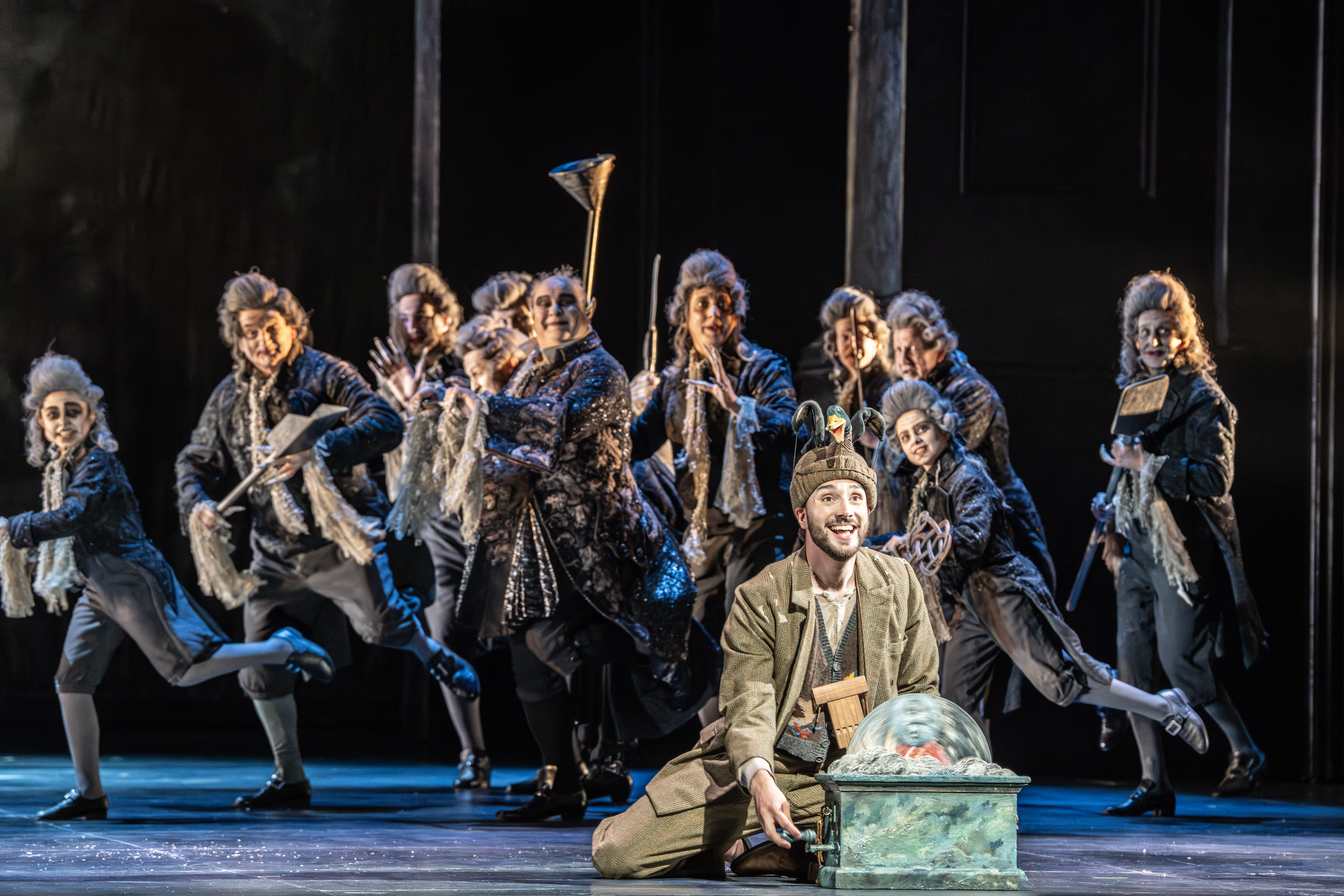 A performer sits on stage holding a domed object. They are flanked by several members of the chorus wearing dress jackets, powder wigs and dark makeup. They are performing in The Royal Opera's production of The Magic Flute. (6 of 7)