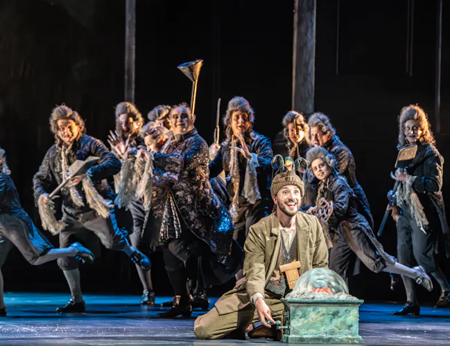 A performer sits on stage holding a domed object. They are flanked by several members of the chorus wearing dress jackets, powder wigs and dark makeup. They are performing in The Royal Opera's production of The Magic Flute. (6 of 7)