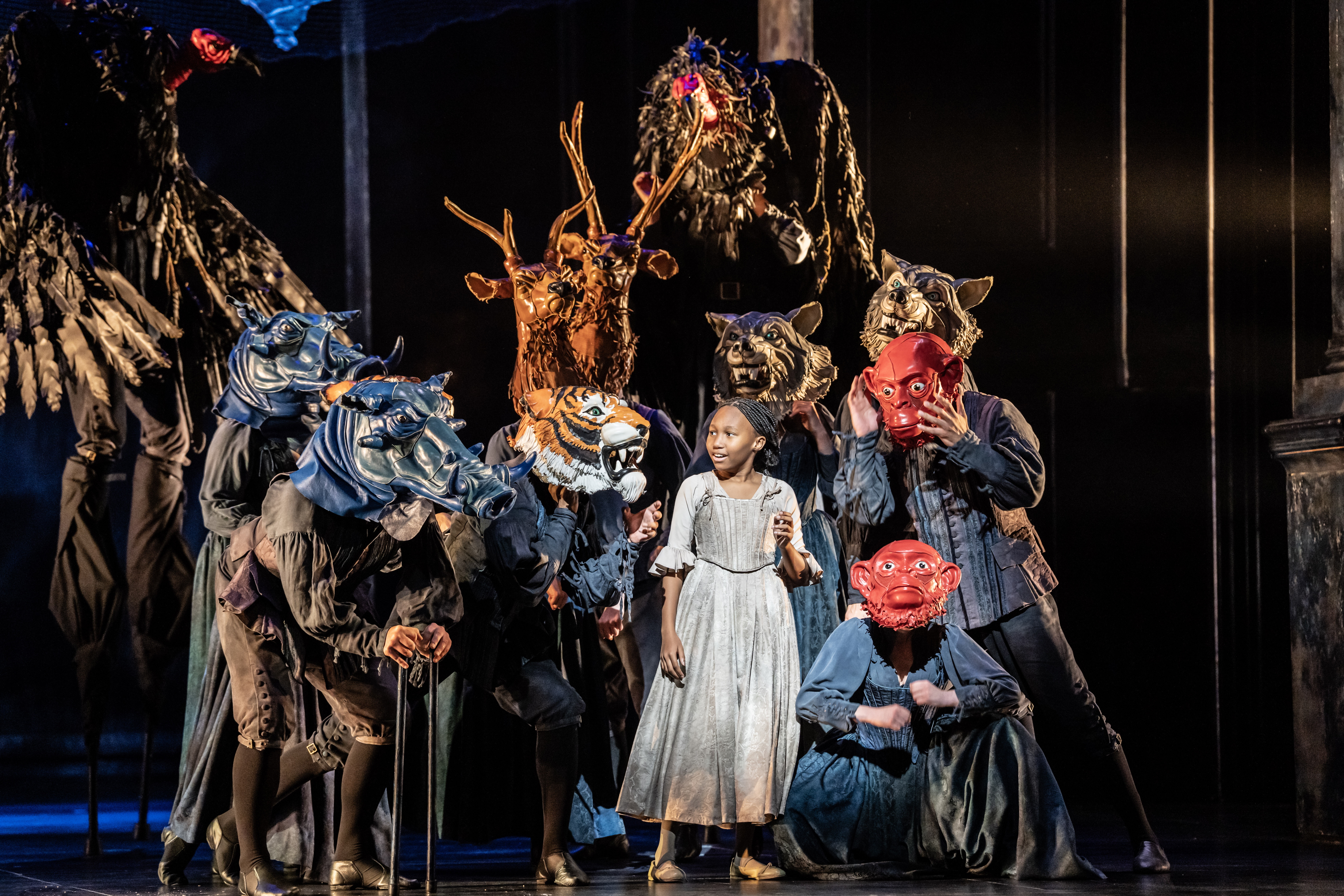 A child performers stands on stage wearing a grey medieval-style dress/. They are flanked by several other performers wearing a variety of animal or monster masks. They are performing in The Royal Opera's production of The Magic Flute. (5 of 7)