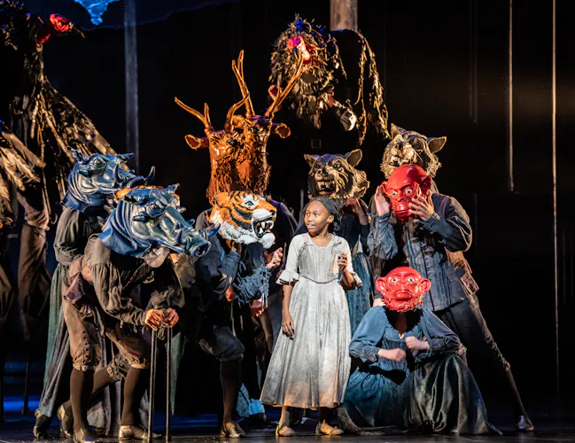 A child performers stands on stage wearing a grey medieval-style dress/. They are flanked by several other performers wearing a variety of animal or monster masks. They are performing in The Royal Opera's production of The Magic Flute. (5 of 7)