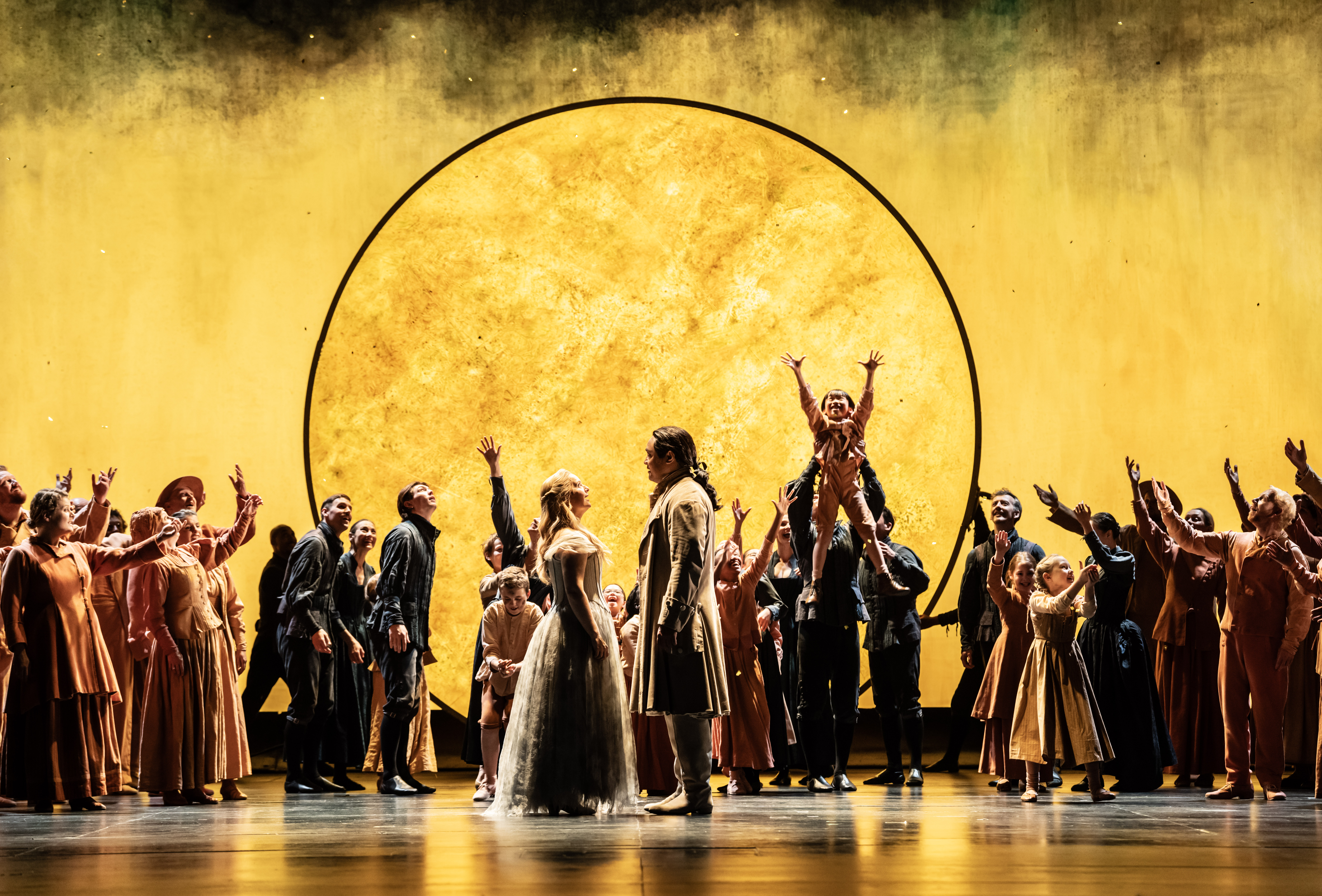 A couple face each other in front of a brightly lit yellow backdrop with the outline of the sun. They are flanked either side by a chorus of performers, one holds up a child to reach the sky. They are performing in The Royal Opera's production of The Magic Flute. (7 of 7)