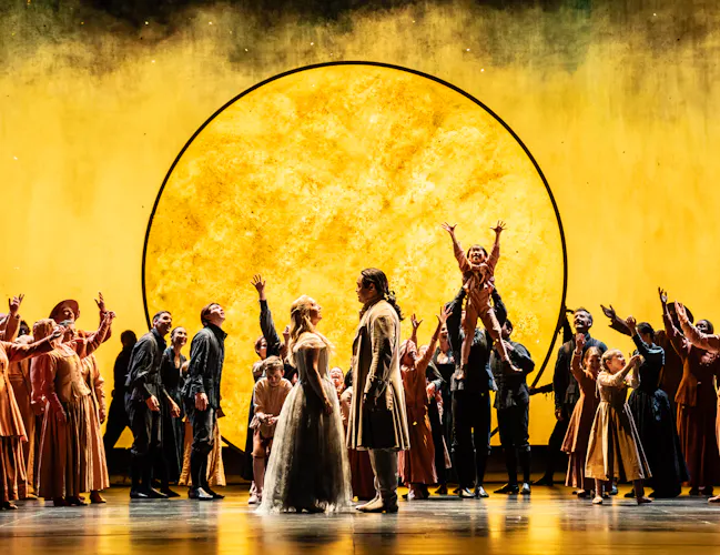 A couple face each other in front of a brightly lit yellow backdrop with the outline of the sun. They are flanked either side by a chorus of performers, one holds up a child to reach the sky. They are performing in The Royal Opera's production of The Magic Flute. (7 of 7)