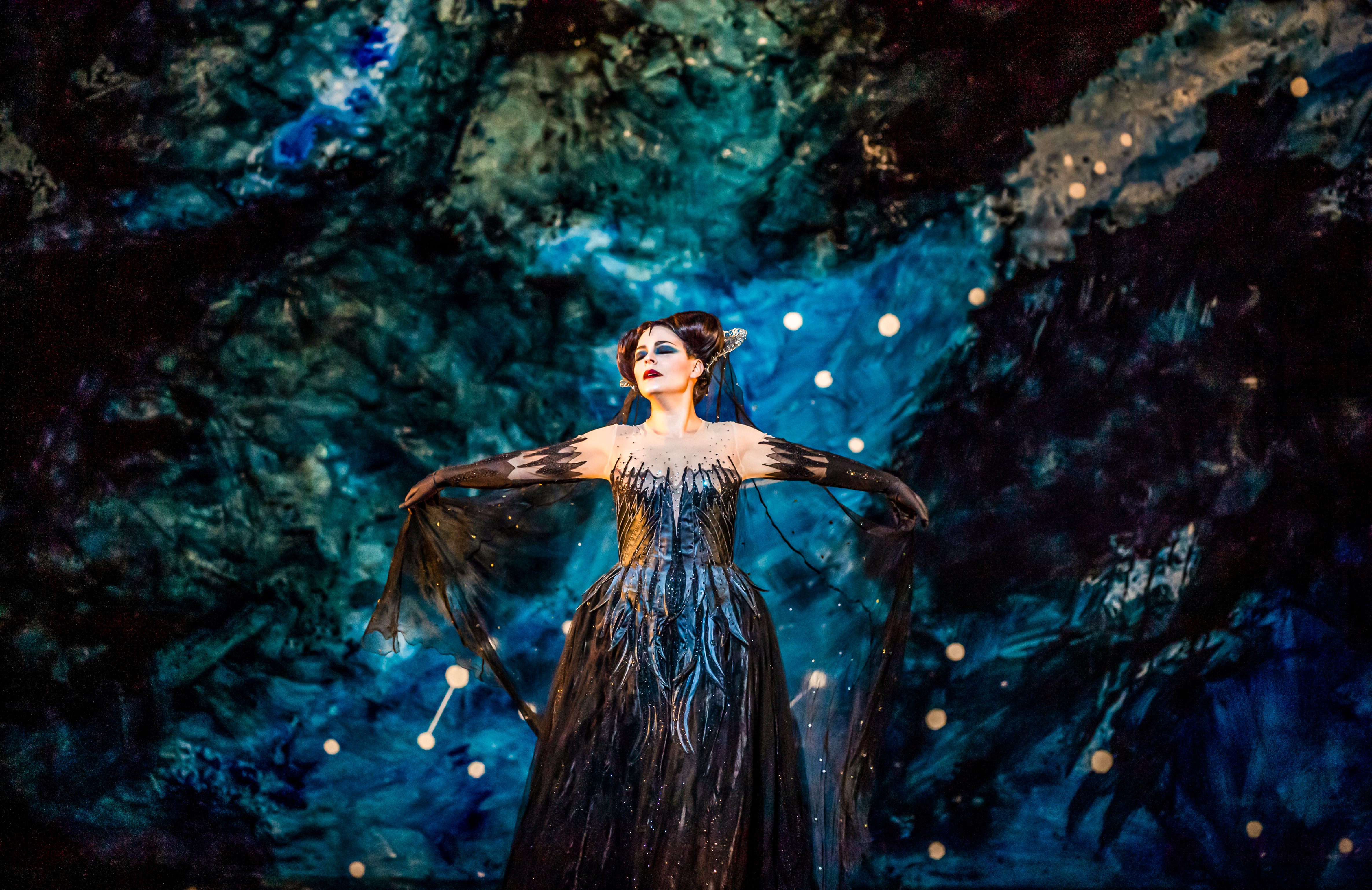 The opera singer Tuuli Takala wears a black ballgown and a medieval style headdress and stands in front of a backdrop painted like an astrological sky. She holds her arms outwards as she sings. She is performing as Queen of the Night in The Royal Opera's production of The Magic Flute. (4 of 12)