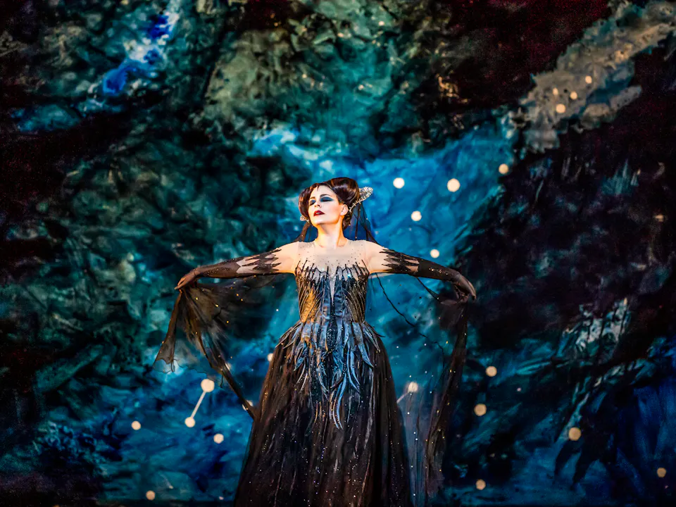 The opera singer Tuuli Takala wears a black ballgown and a medieval style headdress and stands in front of a backdrop painted like an astrological sky. She holds her arms outwards as she sings. She is performing as Queen of the Night in The Royal Opera's production of The Magic Flute.