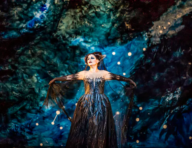 The opera singer Tuuli Takala wears a black ballgown and a medieval style headdress and stands in front of a backdrop painted like an astrological sky. She holds her arms outwards as she sings. She is performing as Queen of the Night in The Royal Opera's production of The Magic Flute. (4 of 12)