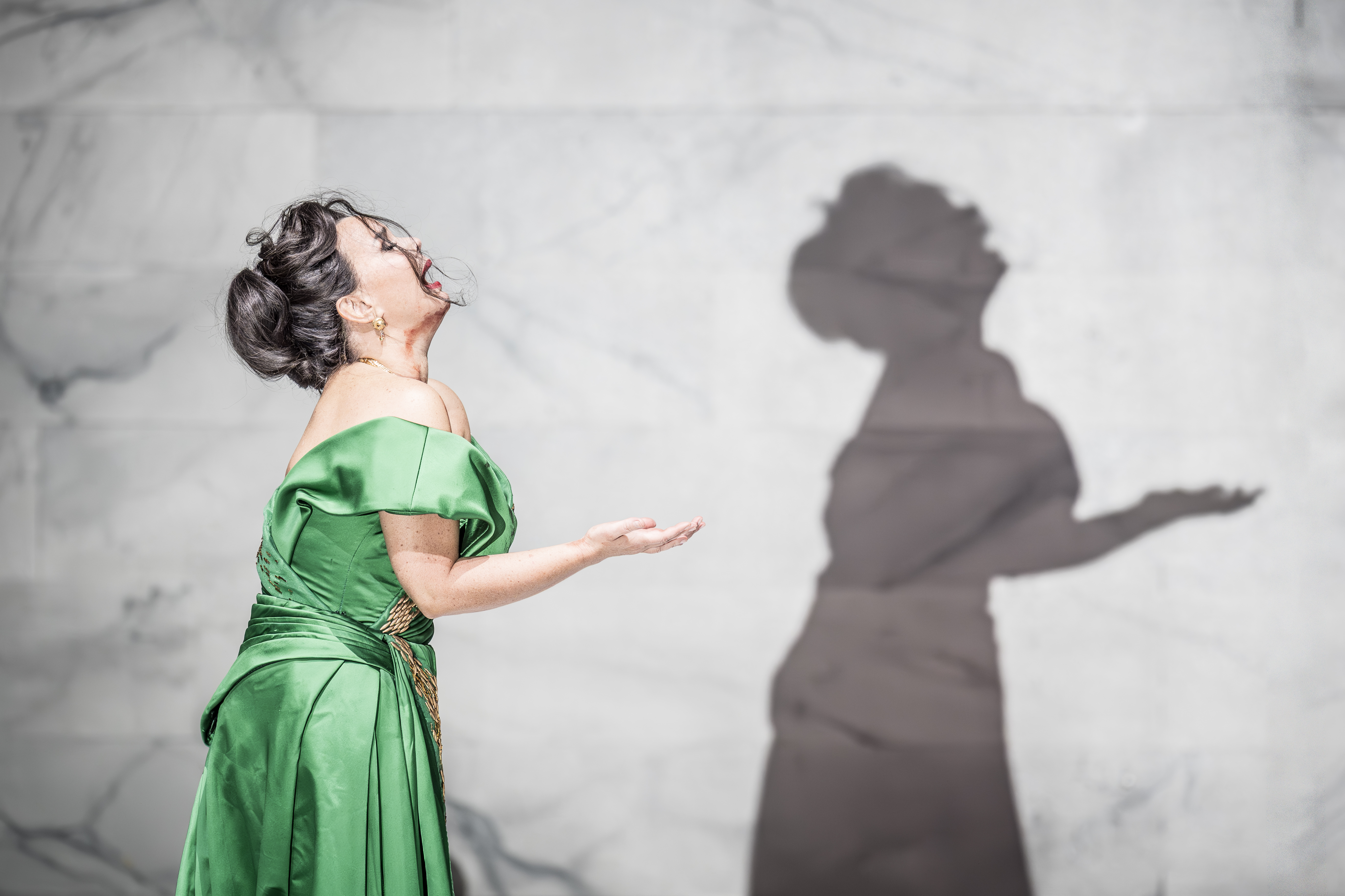 An opera singer wearing a green silk dress sings passionately in front of a backdrop of a marble wall. Her shadow is visible. 