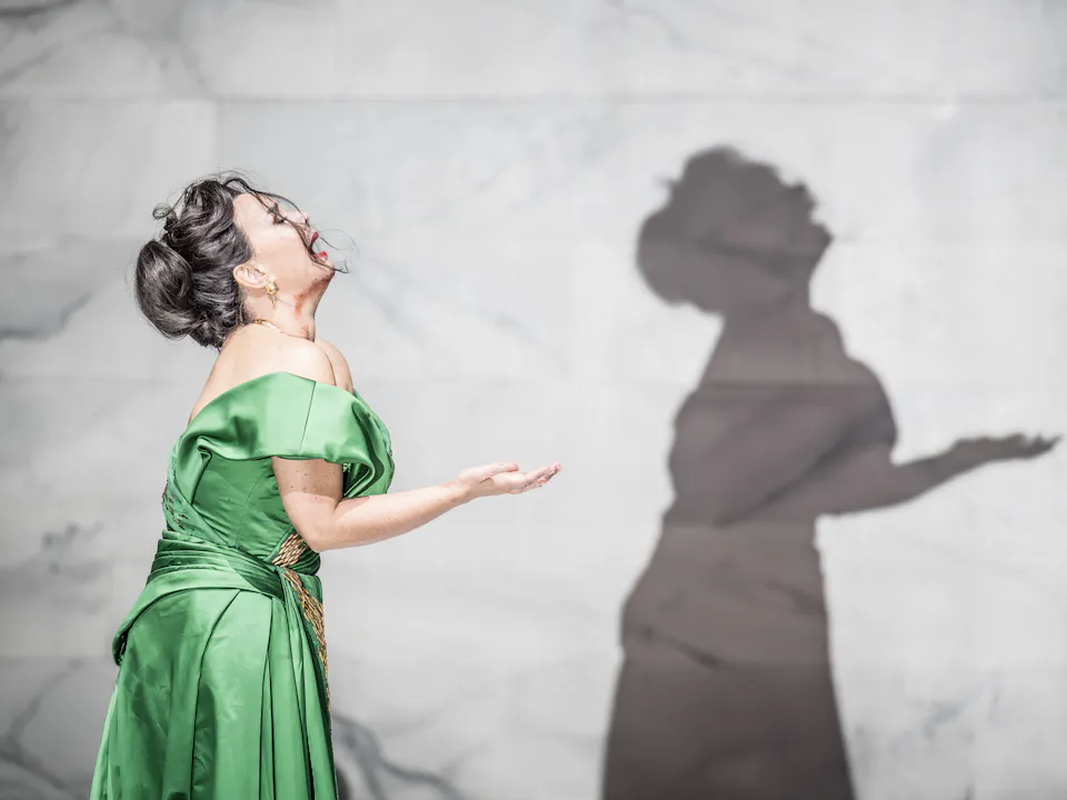 An opera singer wearing a green silk dress sings passionately in front of a backdrop of a marble wall. Her shadow is visible.