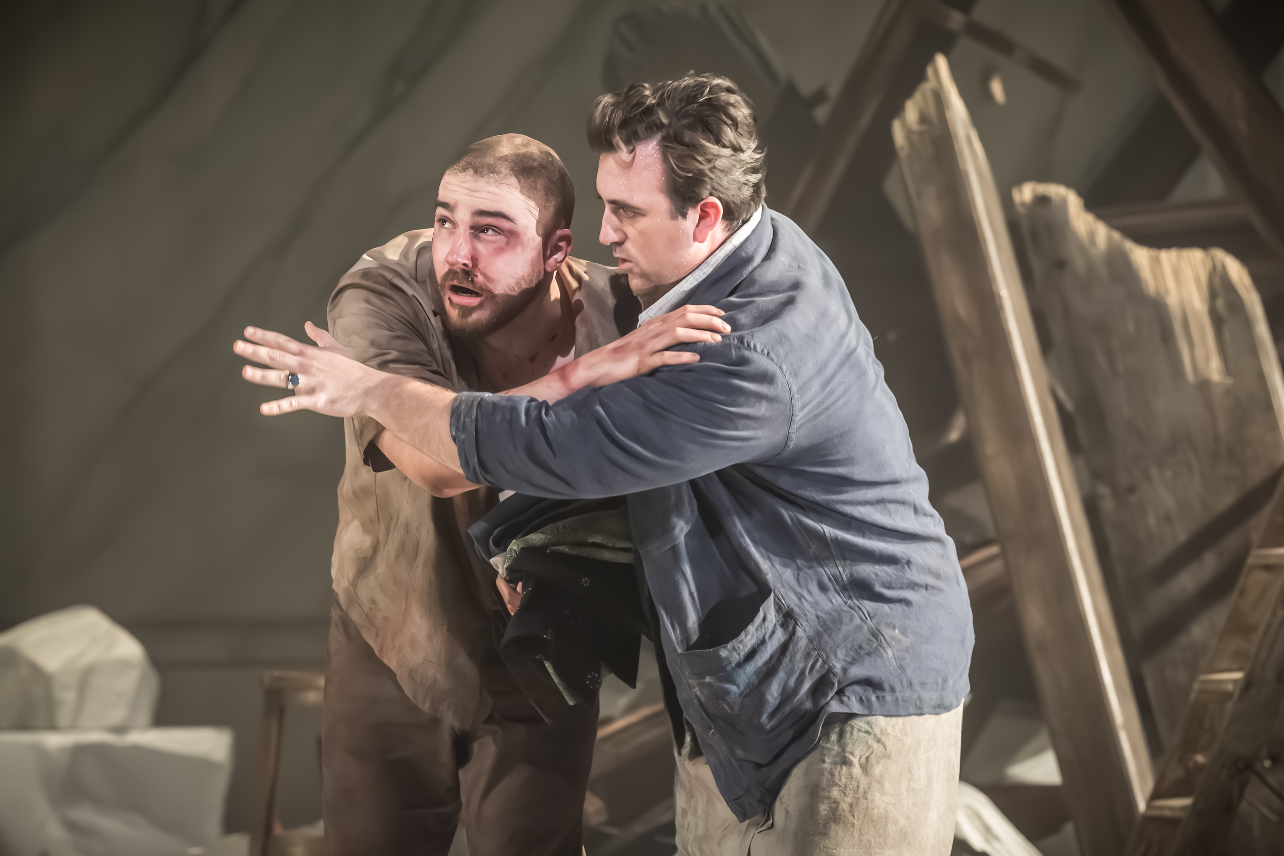 Two performers embrace against a backdrop of wrecked building on stage. One looks as though they have been hurt with bruising and blood on their face. (1 of 6)