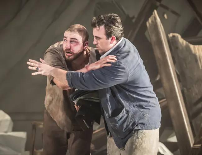 Two performers embrace against a backdrop of wrecked building on stage. One looks as though they have been hurt with bruising and blood on their face. (1 of 6)
