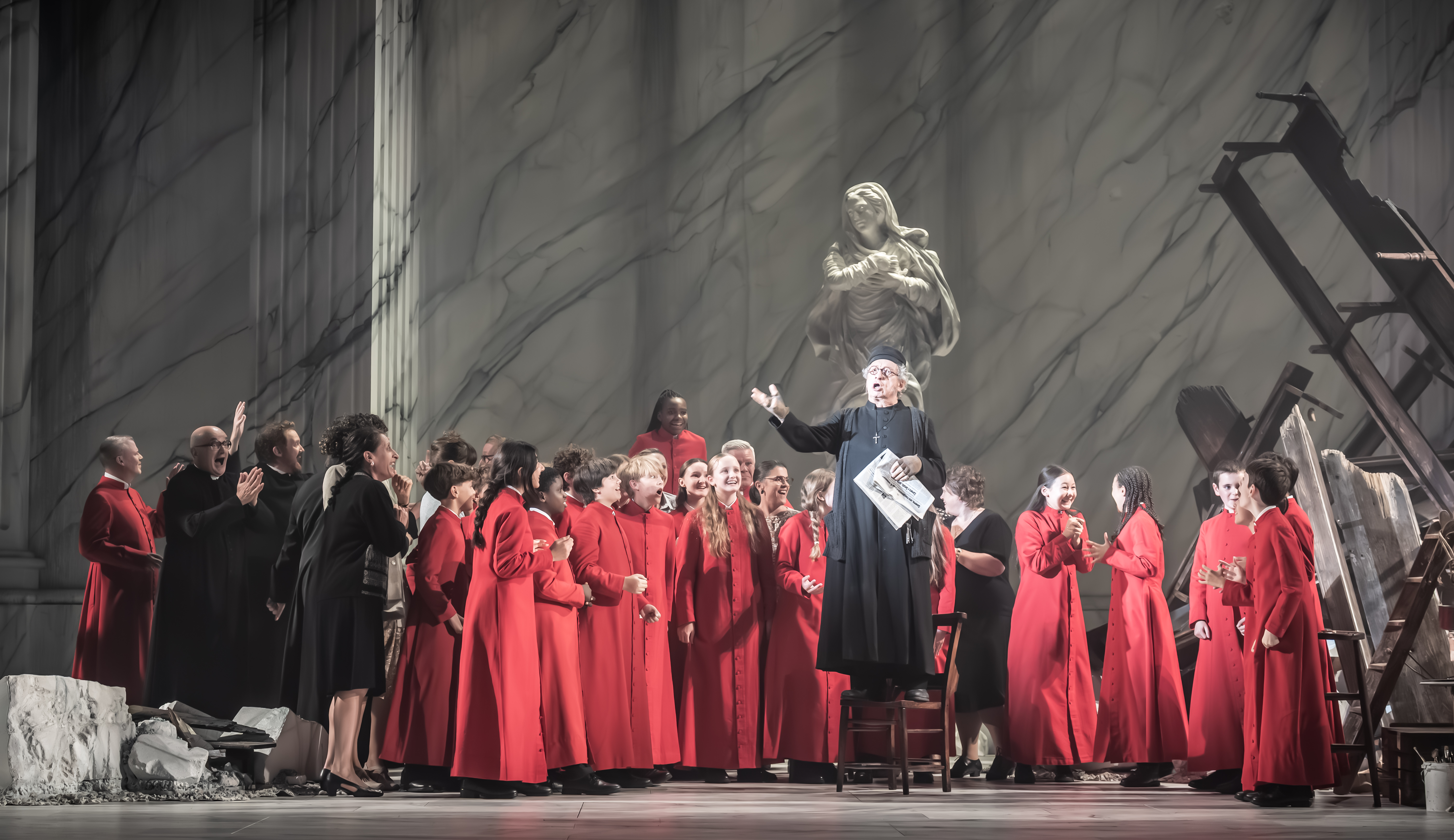 A performer wearing black cleric clothes stands on a chair in the centre of the stage, flanked by many performers in red cleric clothes as well as other performers wearing black. Behind them the stage is set with a wrecked building and a religious statue. (2 of 6)