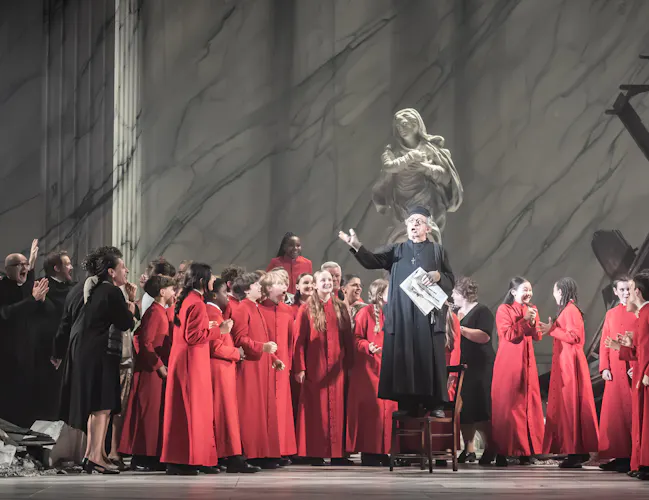 A performer wearing black cleric clothes stands on a chair in the centre of the stage, flanked by many performers in red cleric clothes as well as other performers wearing black. Behind them the stage is set with a wrecked building and a religious statue. (2 of 6)