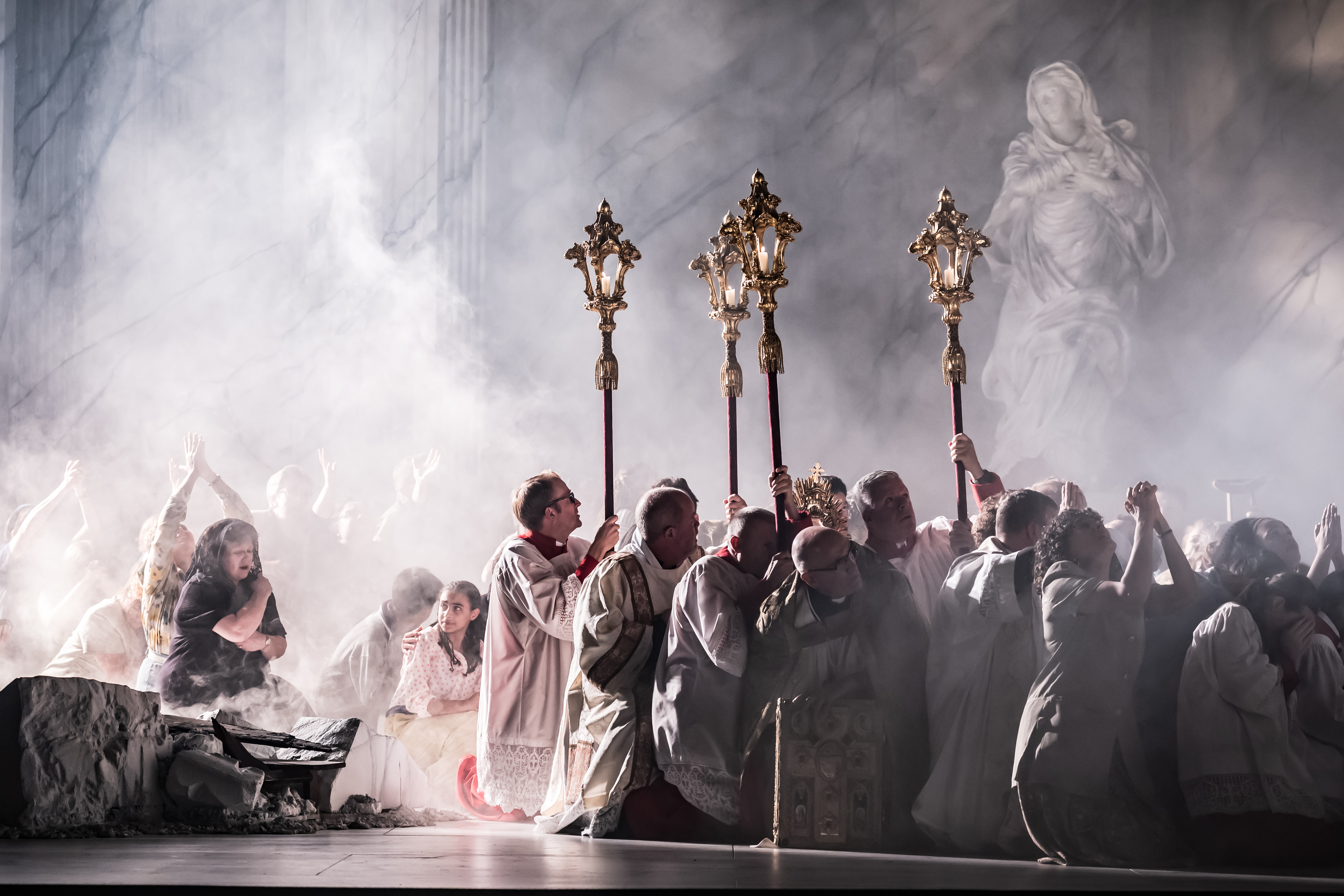 Several performers wearing white vestments crouch on the floor holding up incense thuribles on sticks while other performers dressed as congregation also crouch, as though they are afraid. The stage is set with smoke. (3 of 6)