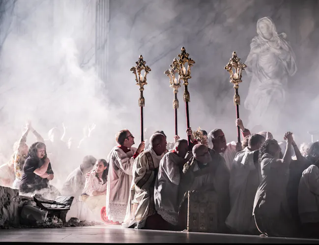 Several performers wearing white vestments crouch on the floor holding up incense thuribles on sticks while other performers dressed as congregation also crouch, as though they are afraid. The stage is set with smoke. (3 of 6)